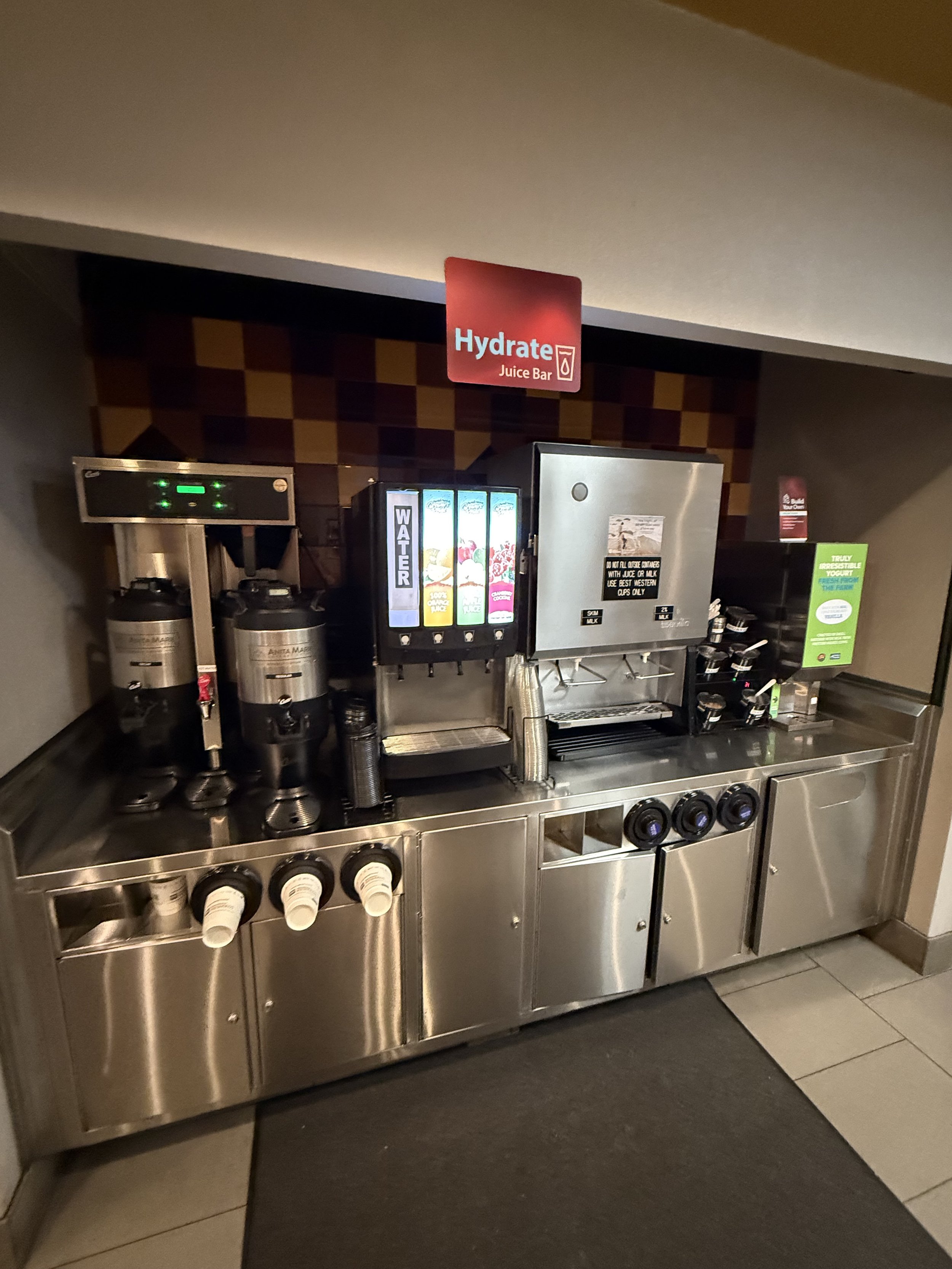 Self-serve beverage station with coffee, juice, and water dispensers, located under a red sign that says 'Hydrate Juice Bar' in a hotel or cafeteria setting.