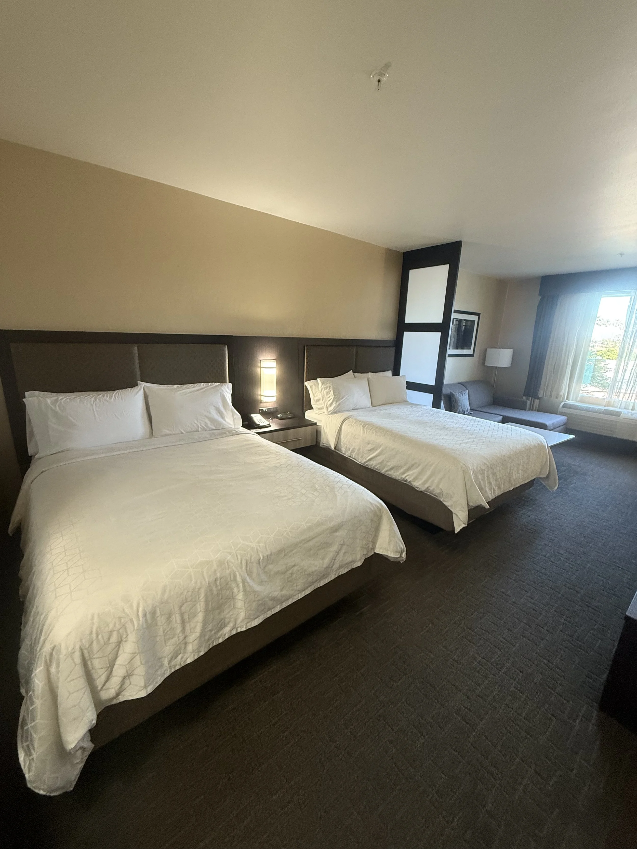 A hotel room with two beds, a nightstand with a phone and lamp between the beds, and a sitting area with a window, curtains, and a sofa.