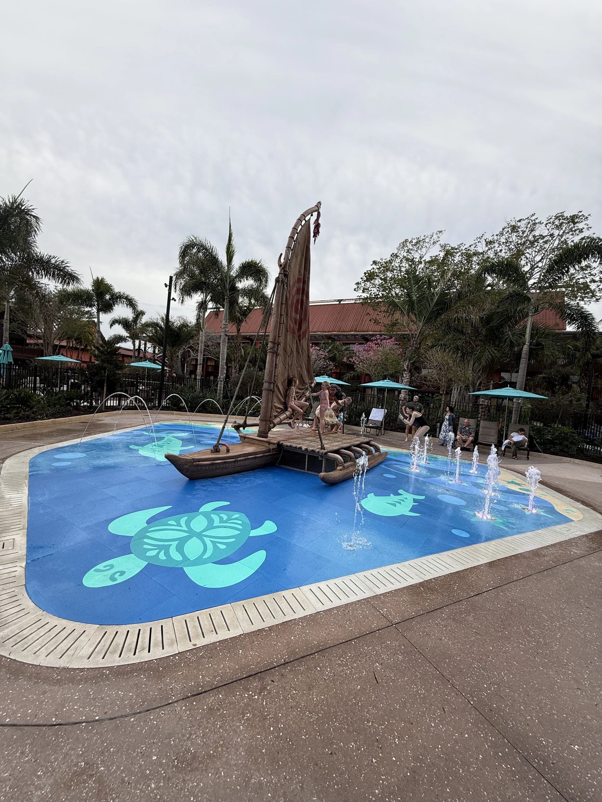 A decorative pool with a pirate ship fountain, water jets, and ocean-themed artwork around the edges, surrounded by palm trees and seating areas.