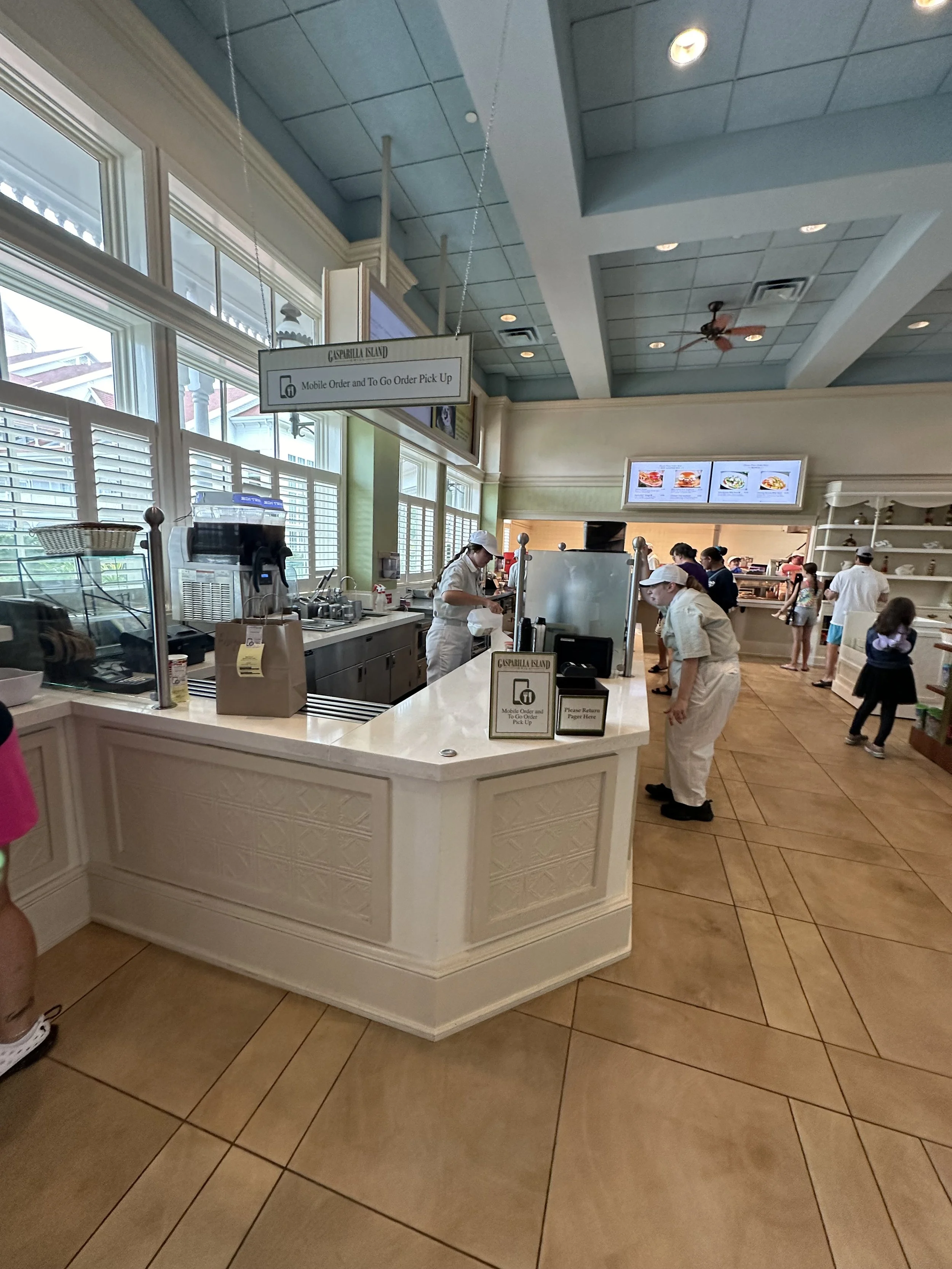 Inside a restaurant with a counter for mobile order pick up, several employees in white uniforms working, large windows allowing natural light, digital menu screens on the wall, customers waiting or placing orders, and a sign indicating mobile order 