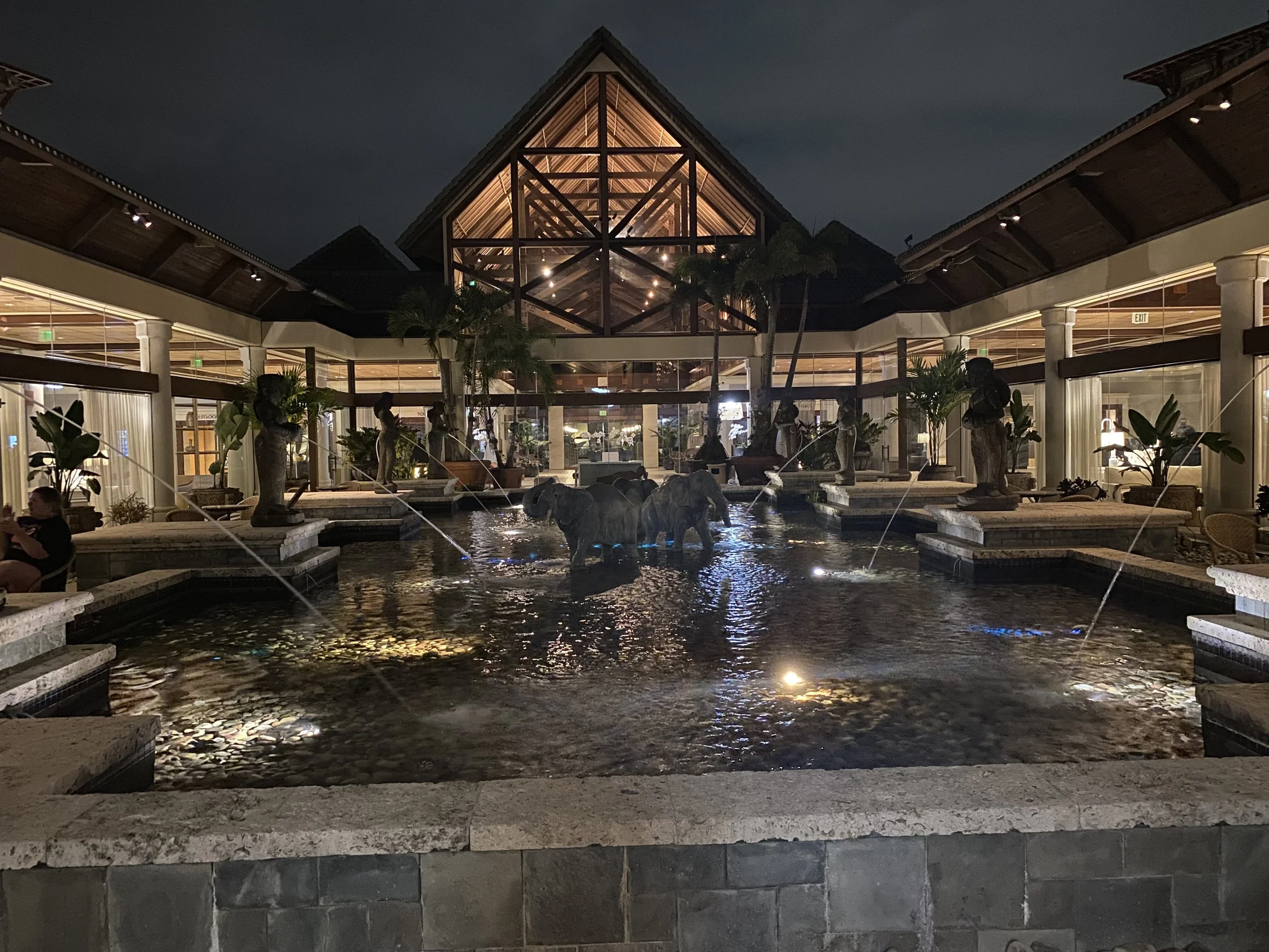 Night view of an indoor hotel lobby featuring a decorative fountain with elephant sculptures and water spouts, surrounded by tropical plants and seating areas, with a high ceiling and wooden beams.