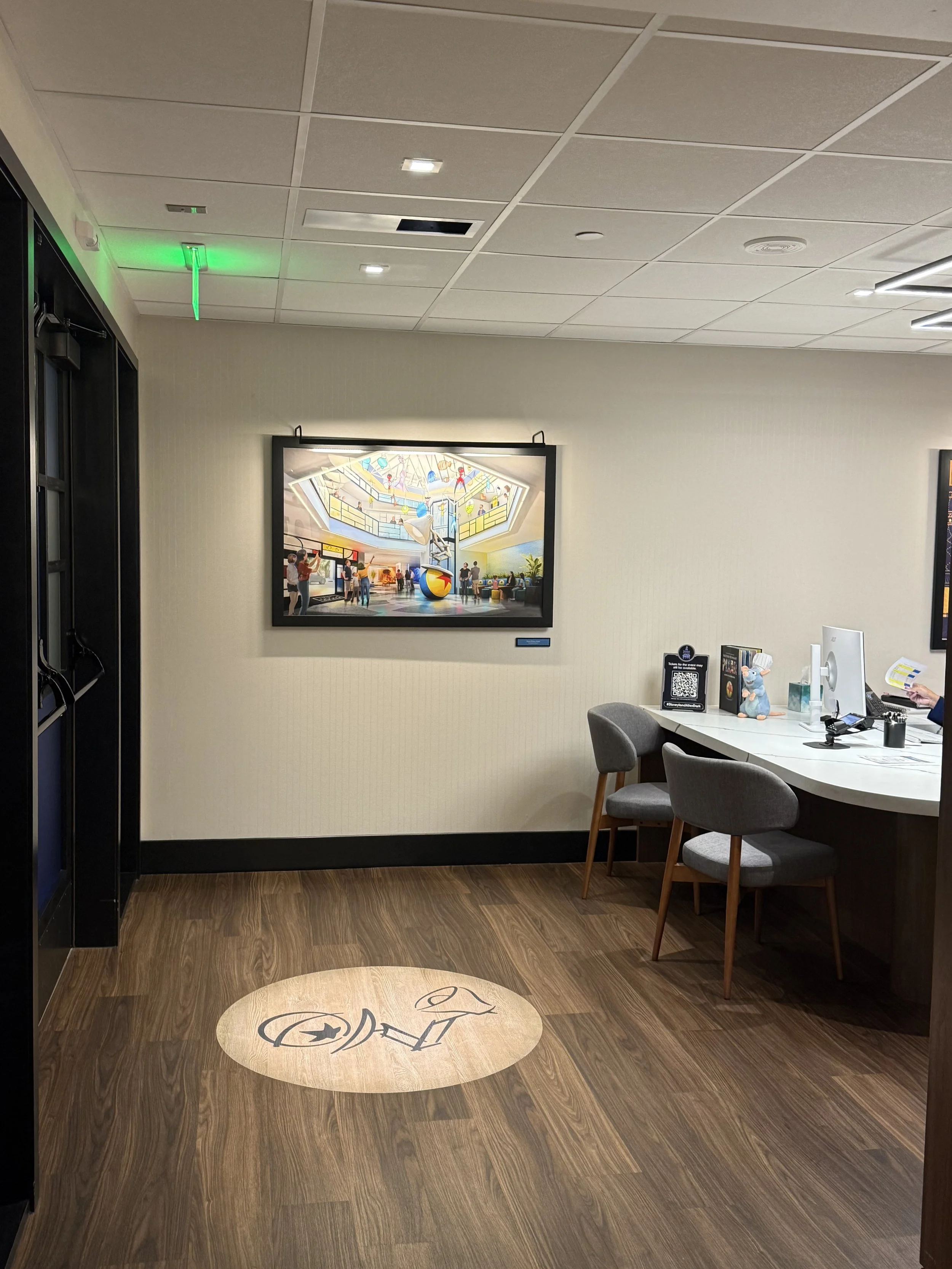 Interior of a modern office or reception area with a wooden floor, gray chairs, a white desk with a computer, and framed artwork on the wall depicting a busy shopping mall with a large ball