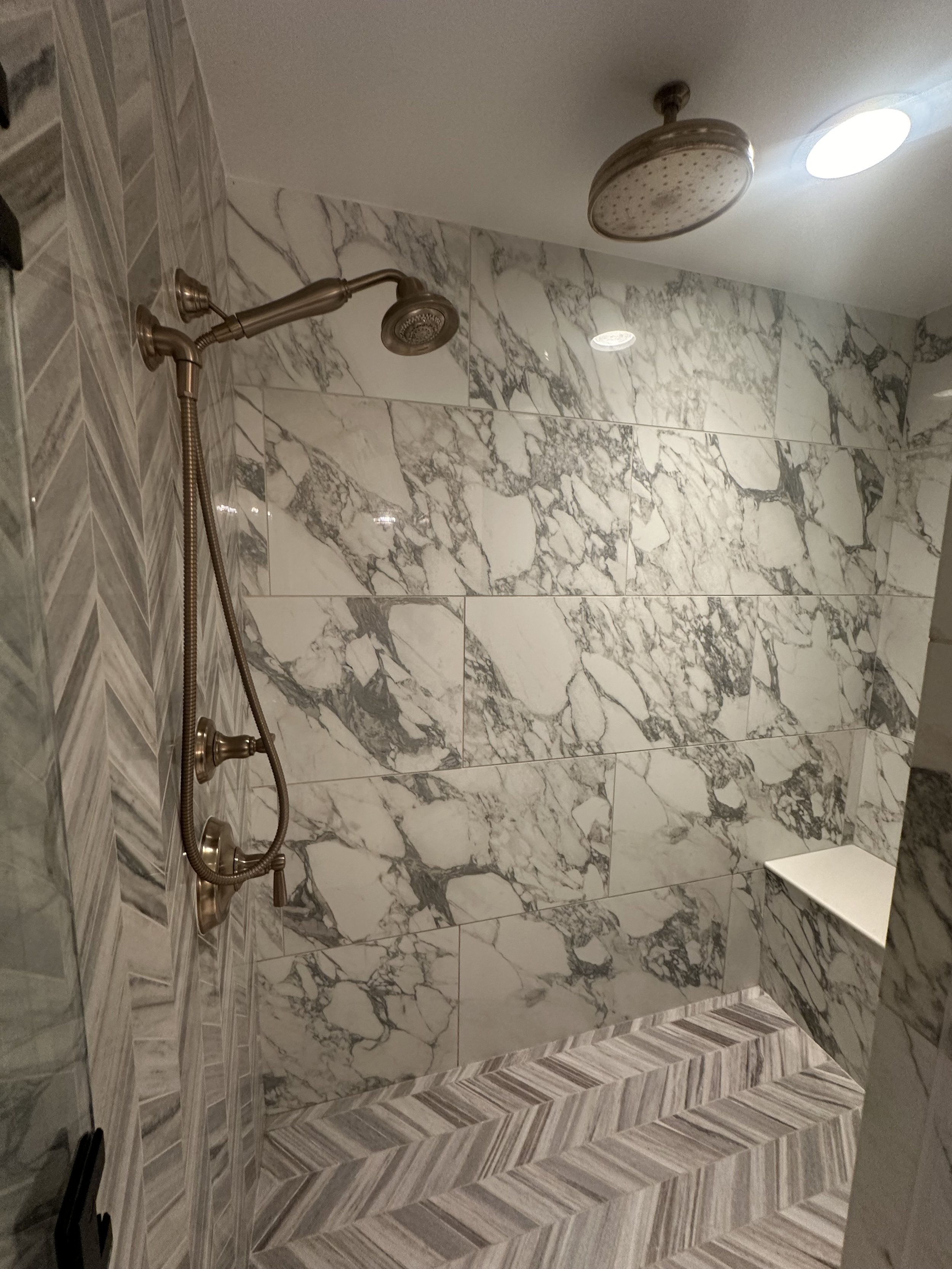 Marble tiled walk-in shower with a overhead rainfall showerhead and a handheld showerhead, and built-in marble shelf in the corner for toiletries.