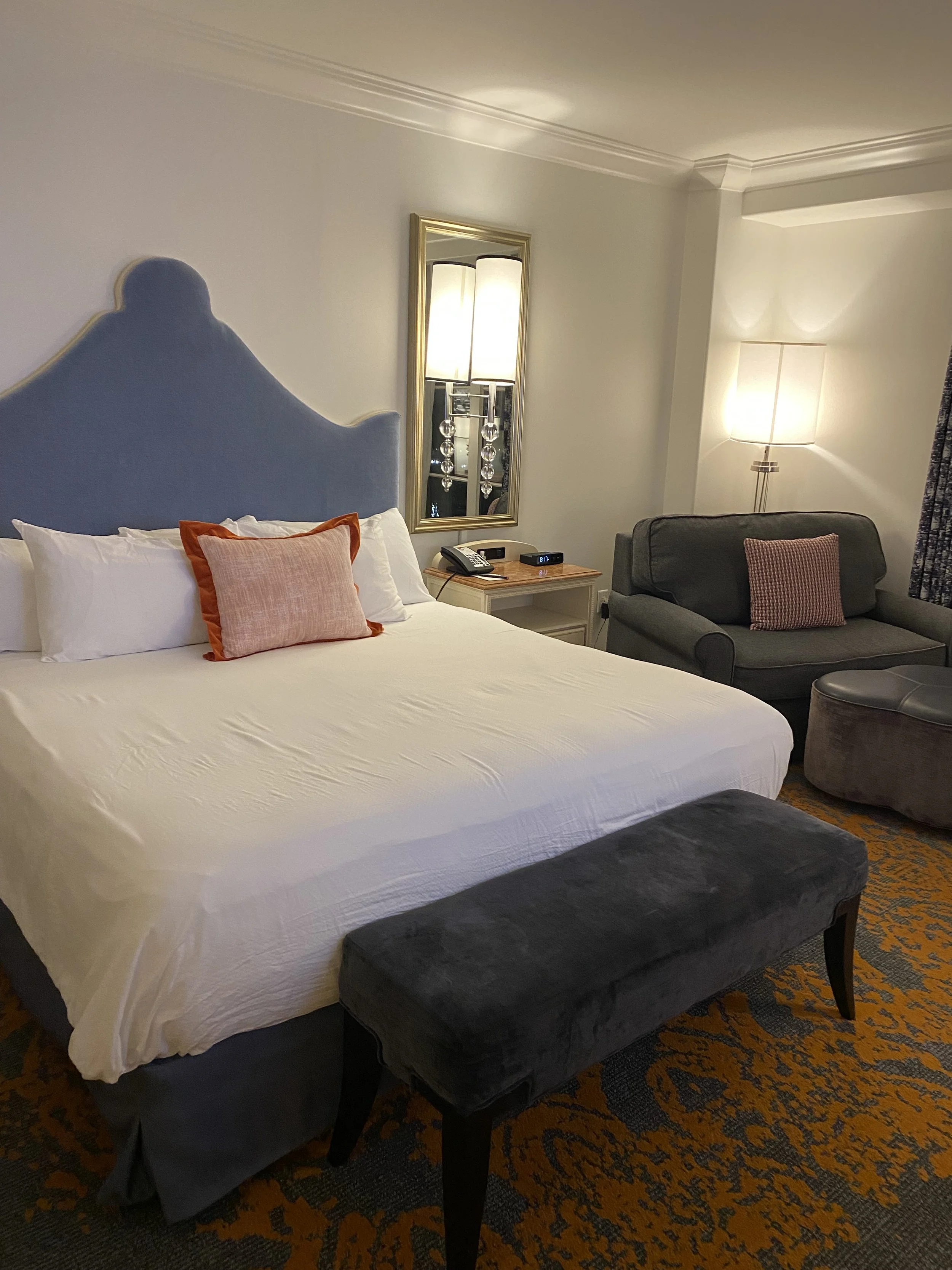Hotel room with a king-sized bed, blue headboard, white bedding, and orange accent pillow. A gray bench at the foot of the bed. To the right, a sitting area with a gray couch, patterned pillow, and a floor lamp. A mirror and a nightstand with a telep