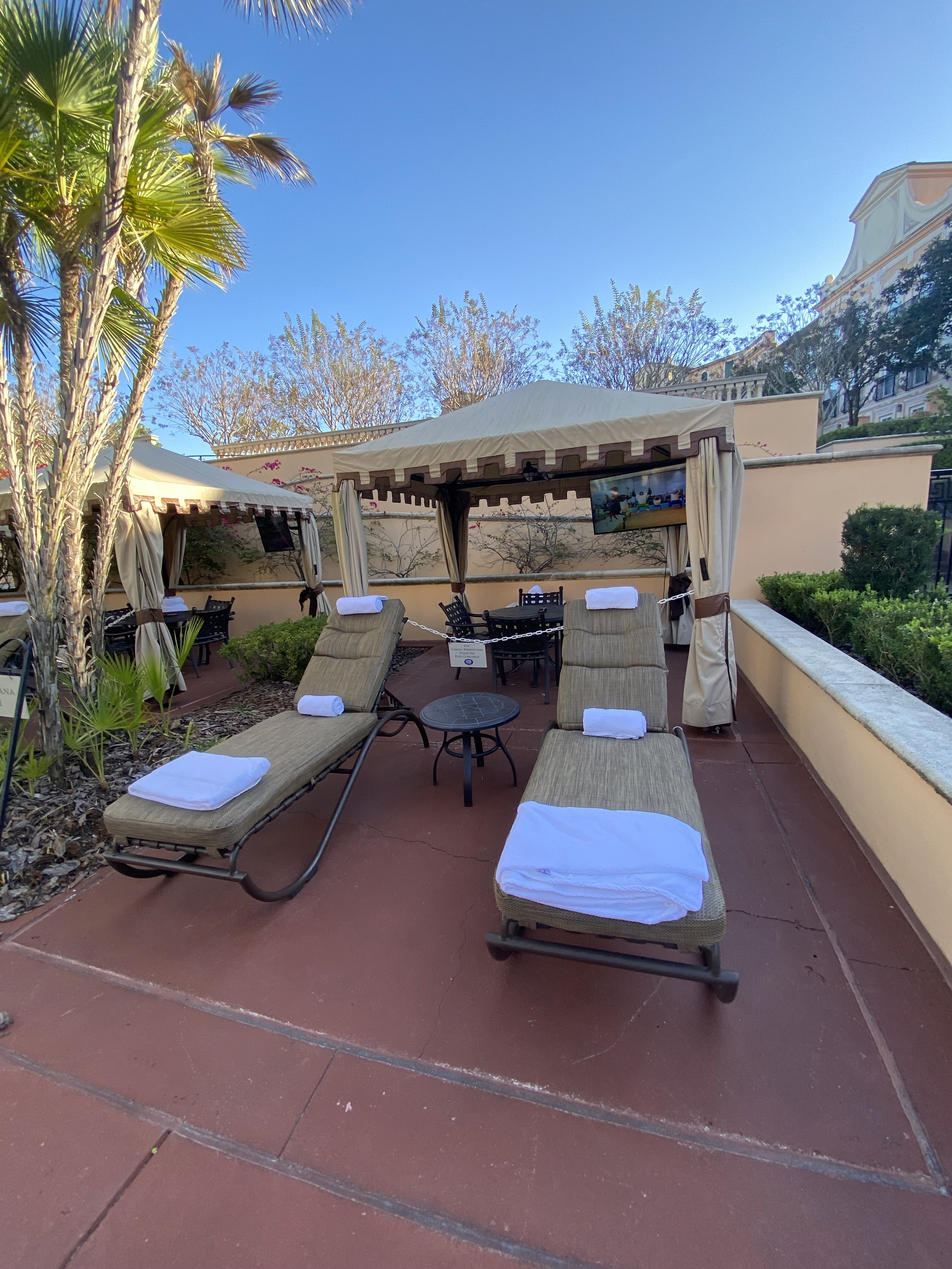 Outdoor patio with lounge chairs, towels, private cabanas, small tables, and a TV, under a clear blue sky.