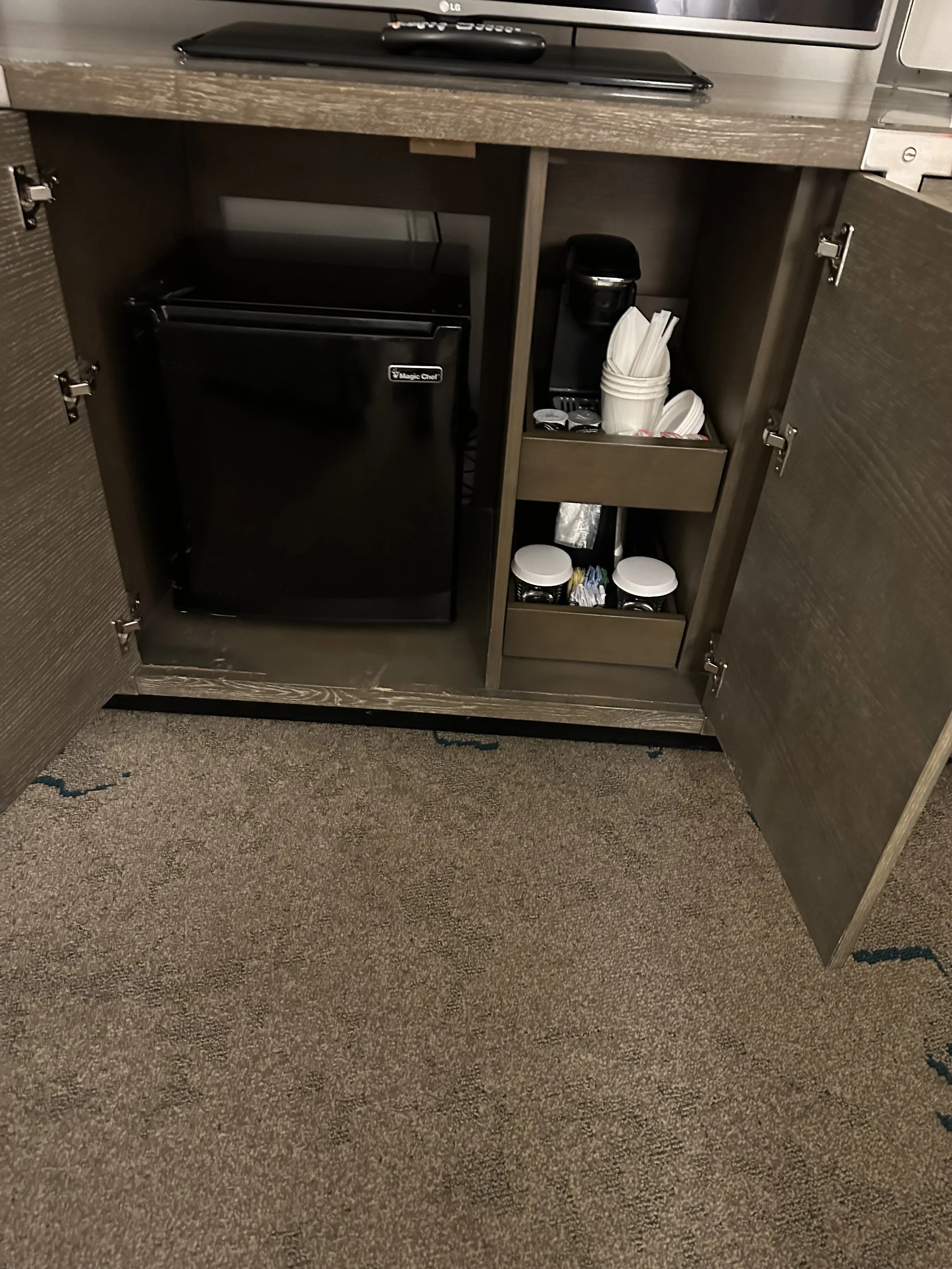 A hotel room kitchenette with a mini fridge, coffee maker, and a tray of cups and condiments inside an open cabinet.