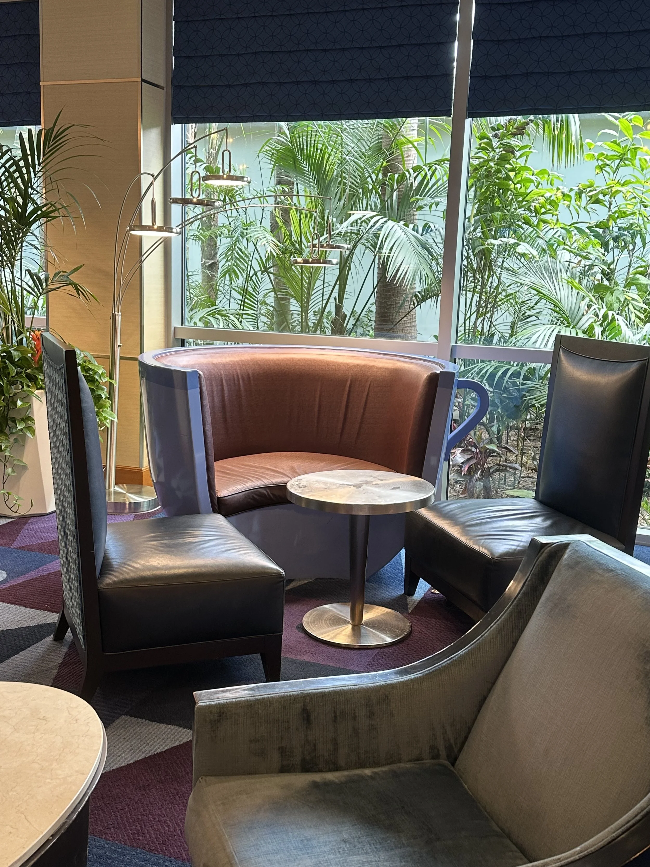 A cozy seating area with various chairs and a small round table with a marble top. Behind the seating area, there is a large window with lush green plants outside. The room has modern decor with a colorful carpet and an arched floor lamp.