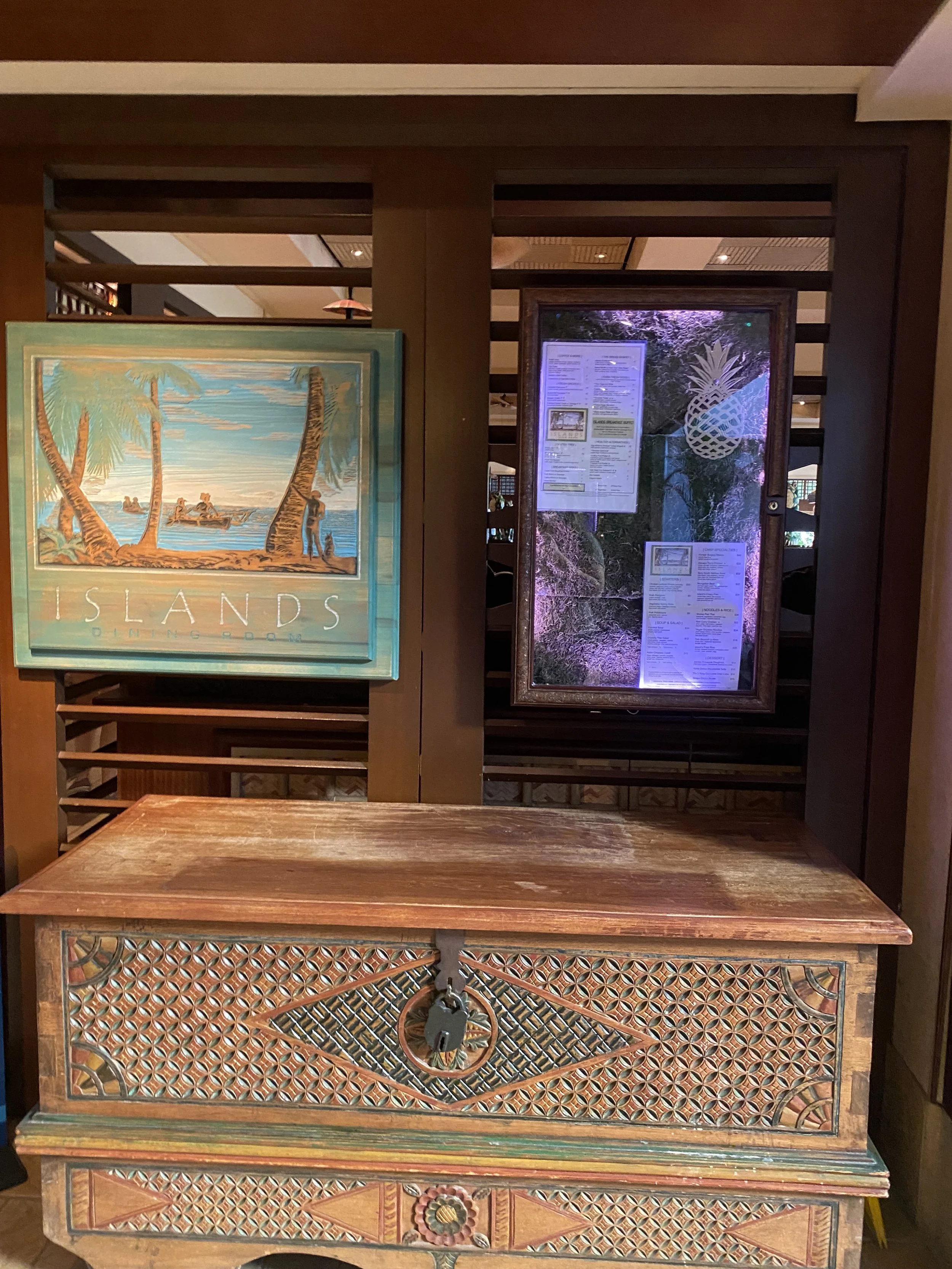 A wooden reception desk in front of a wooden partition. Two framed signs and a menu are hung on the partition, with a painting of a tropical scene on the left.