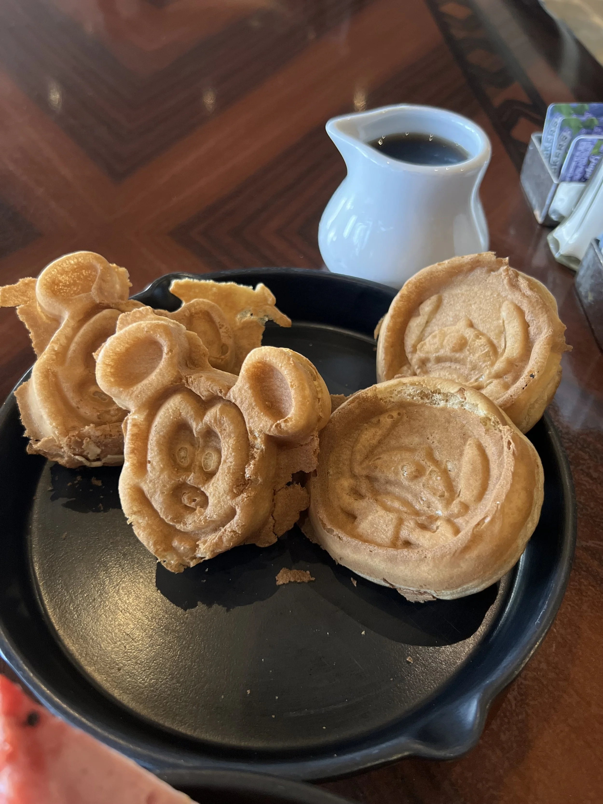 Four Mickey Mouse-shaped waffles on a black plate with a small pitcher of syrup and a table with sugar and cream packets.