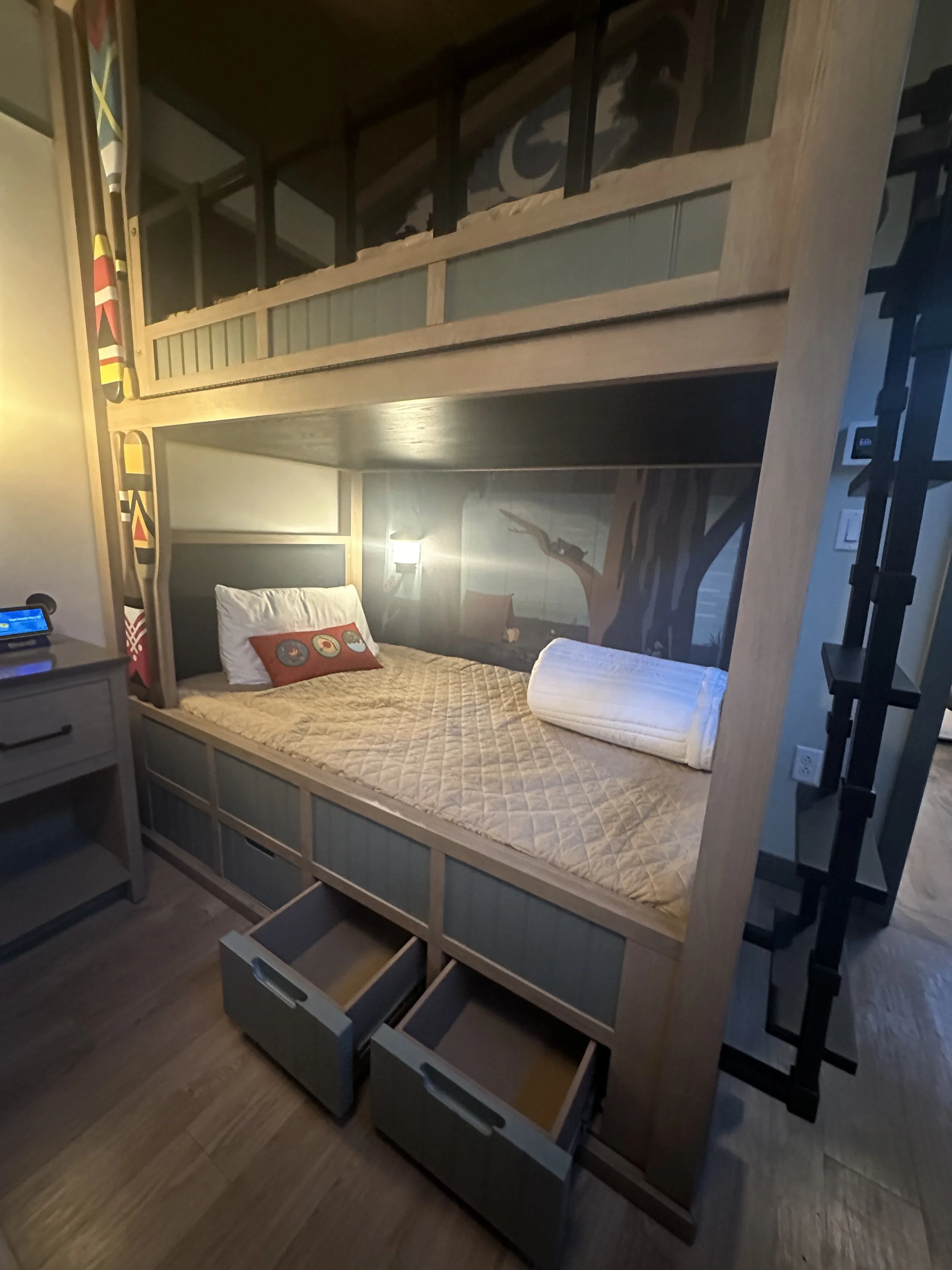 A bedroom with a bunk bed, featuring a lower bed with a beige quilt, a white pillow with an orange pillowcase, and a rolled white towel. The upper bunk has a wooden frame and a railing. There's a wall-mounted lamp above the lower bed, a small nightst