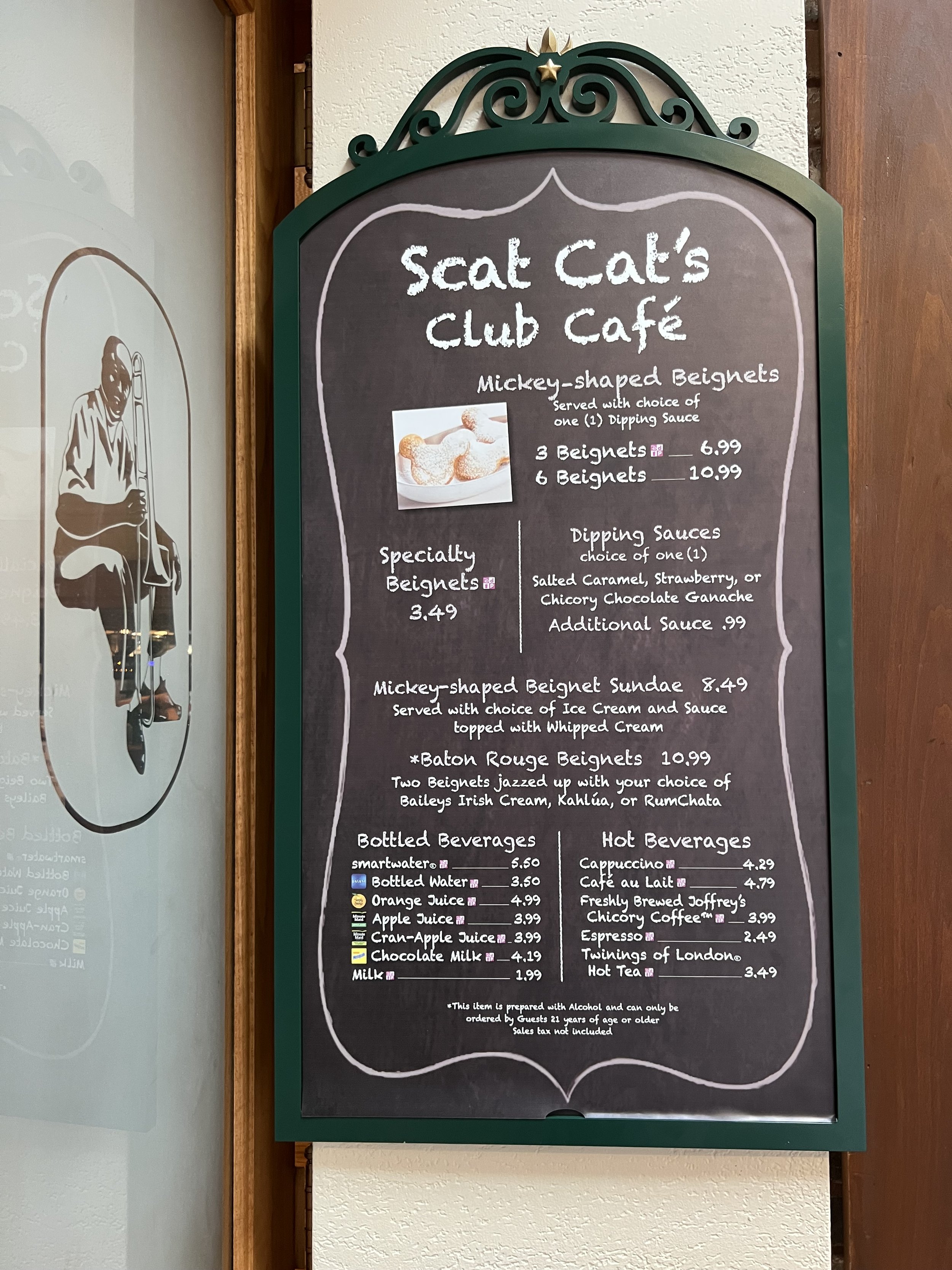 The blackboard menu features a selection of beignets, including Mickey-shaped, specialty, and bourbon varieties, along with bottled and hot beverages at a cafe named 'Scat Cat's Club Café'.