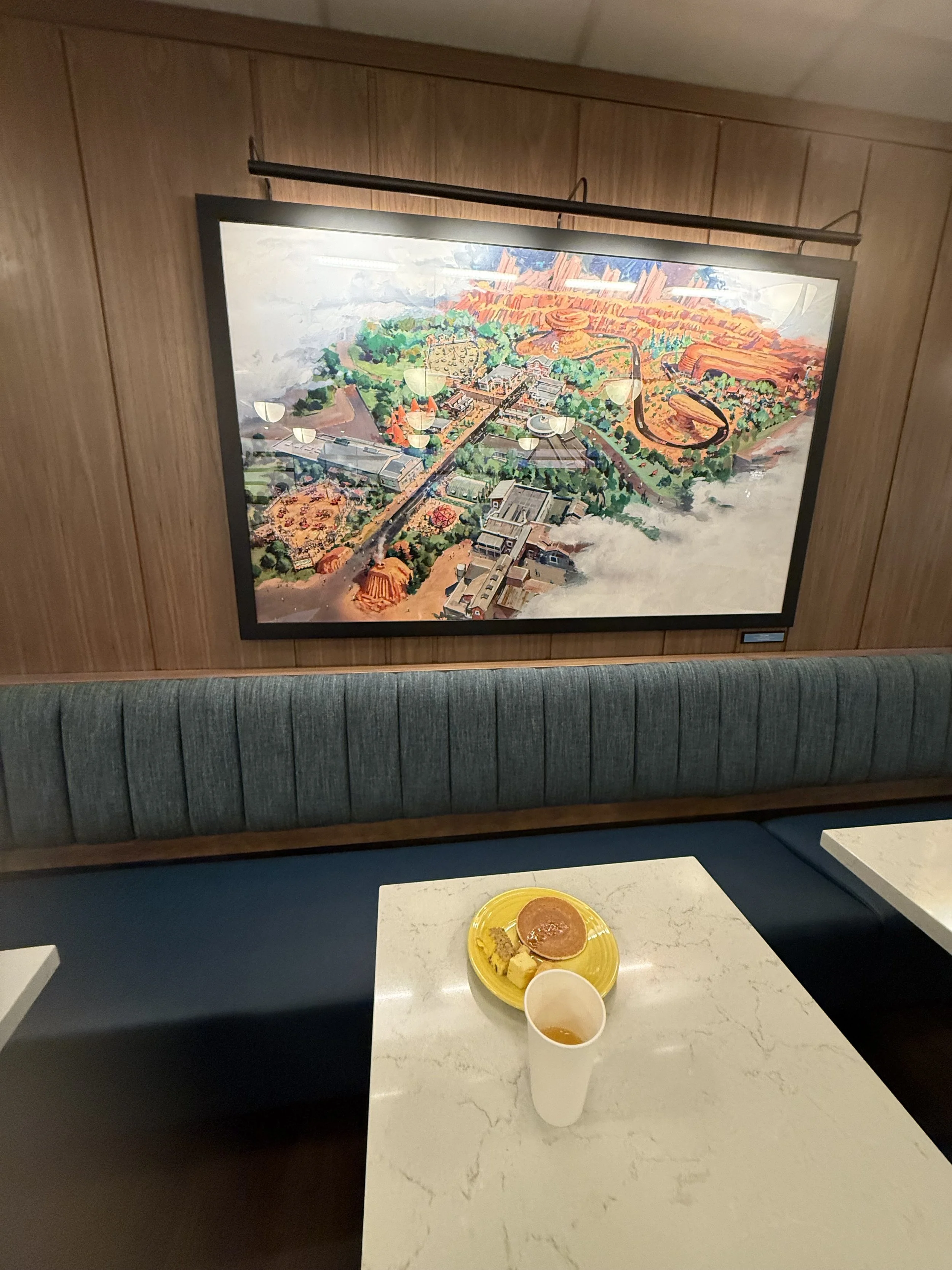 A wall-mounted framed map of Disney California Adventure Park above a booth with a blue upholstered backrest. On the table, there is a white cup with a drink and a yellow plate with a cookie and a small snack.