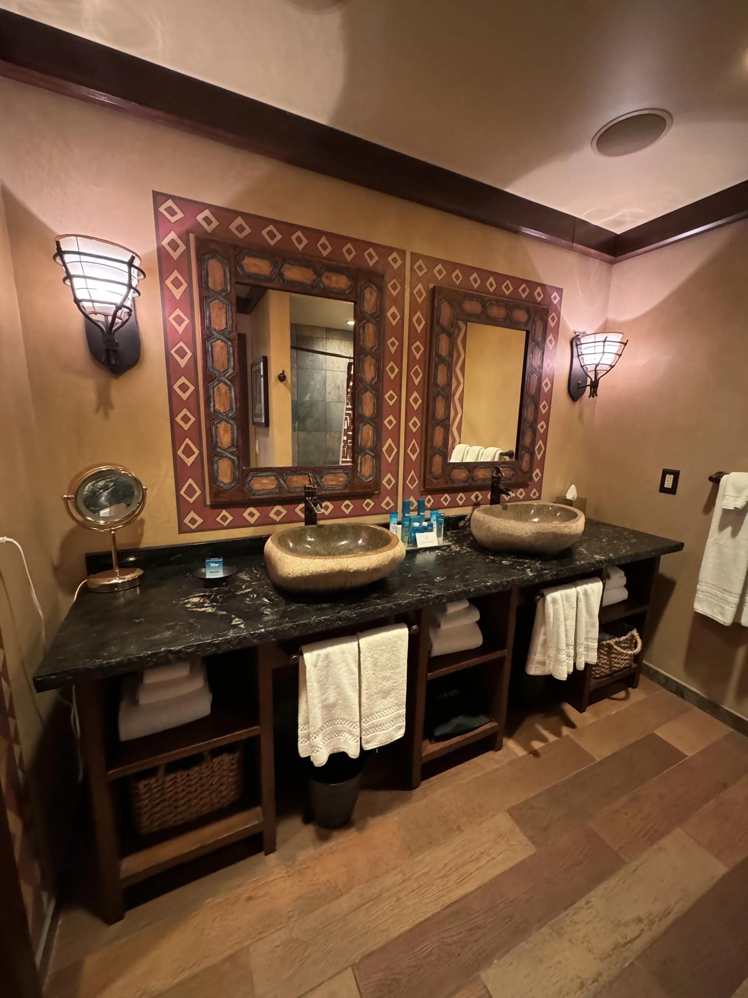 Double vanity bathroom with two stone sinks, large mirrors with decorative frames, wall-mounted lights, and a wooden cabinet with towels underneath.