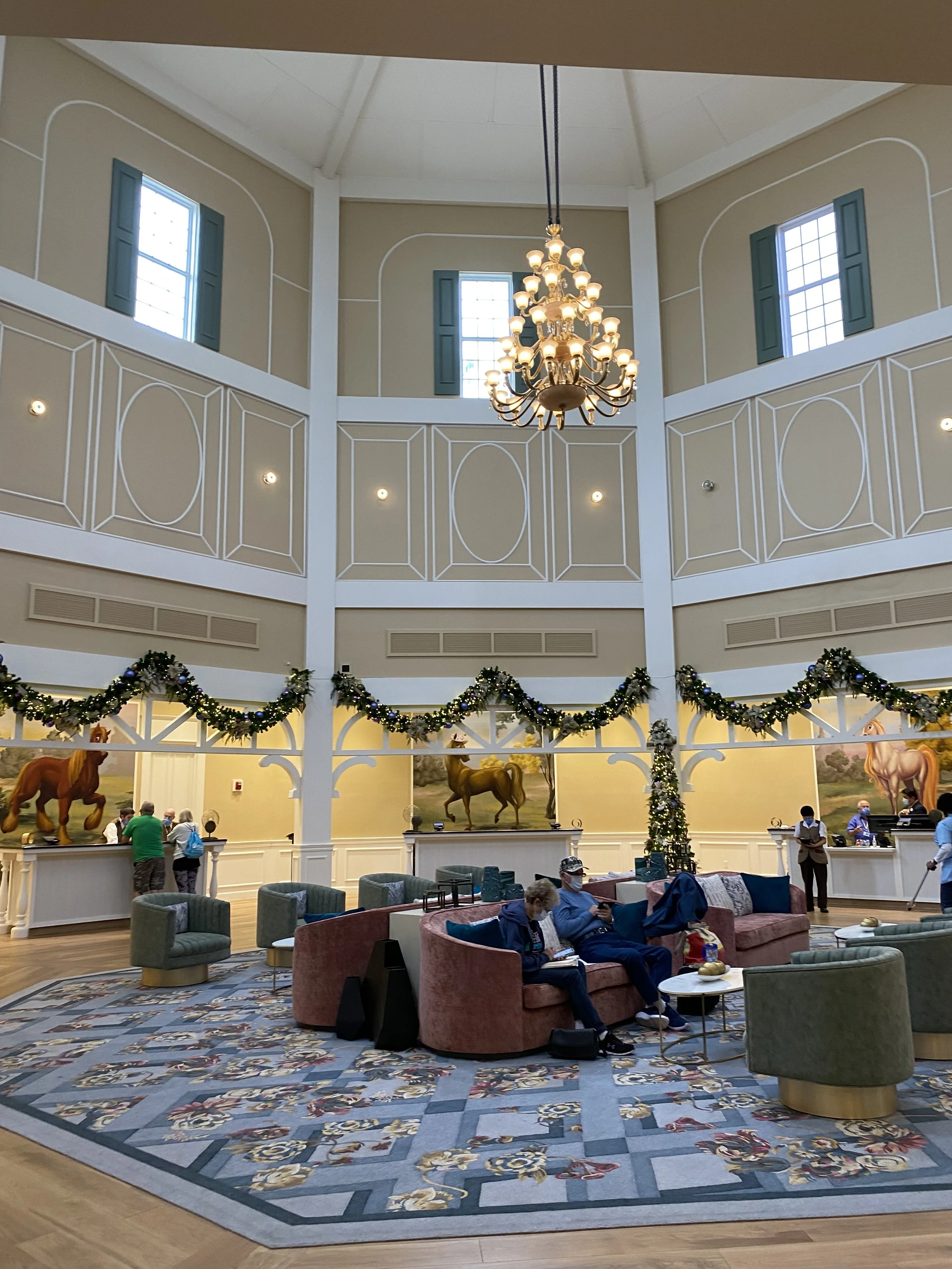 A spacious hotel lobby decorated for Christmas, with a large chandelier, a wreath on a post, and paintings of horses on the wall. Guests are sitting and standing, some wearing masks, with sofas, armchairs, and a patterned rug.