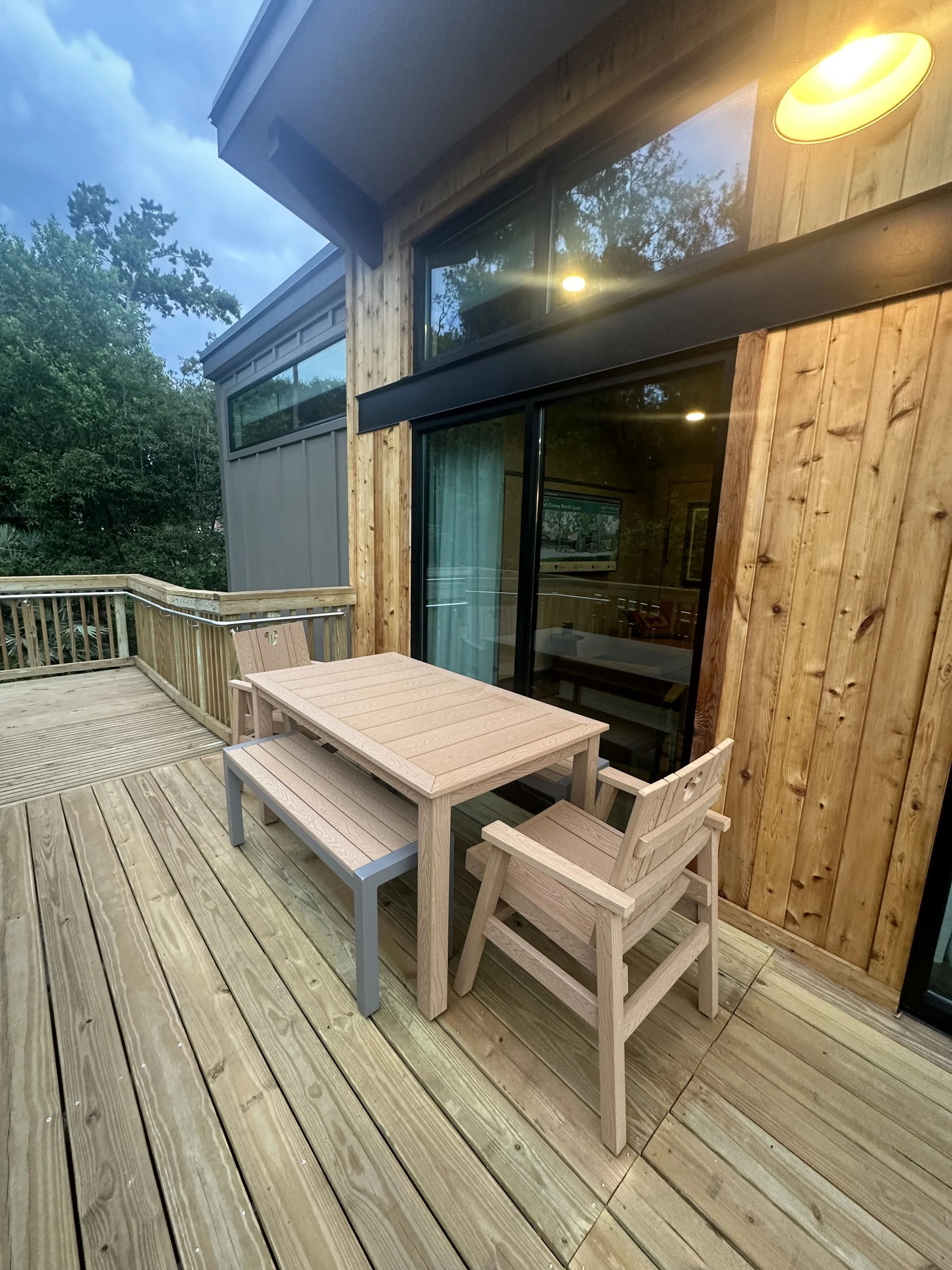 Wooden patio with outdoor table and chairs on a deck, adjacent to a modern house with large glass windows and wooden panel exterior.
