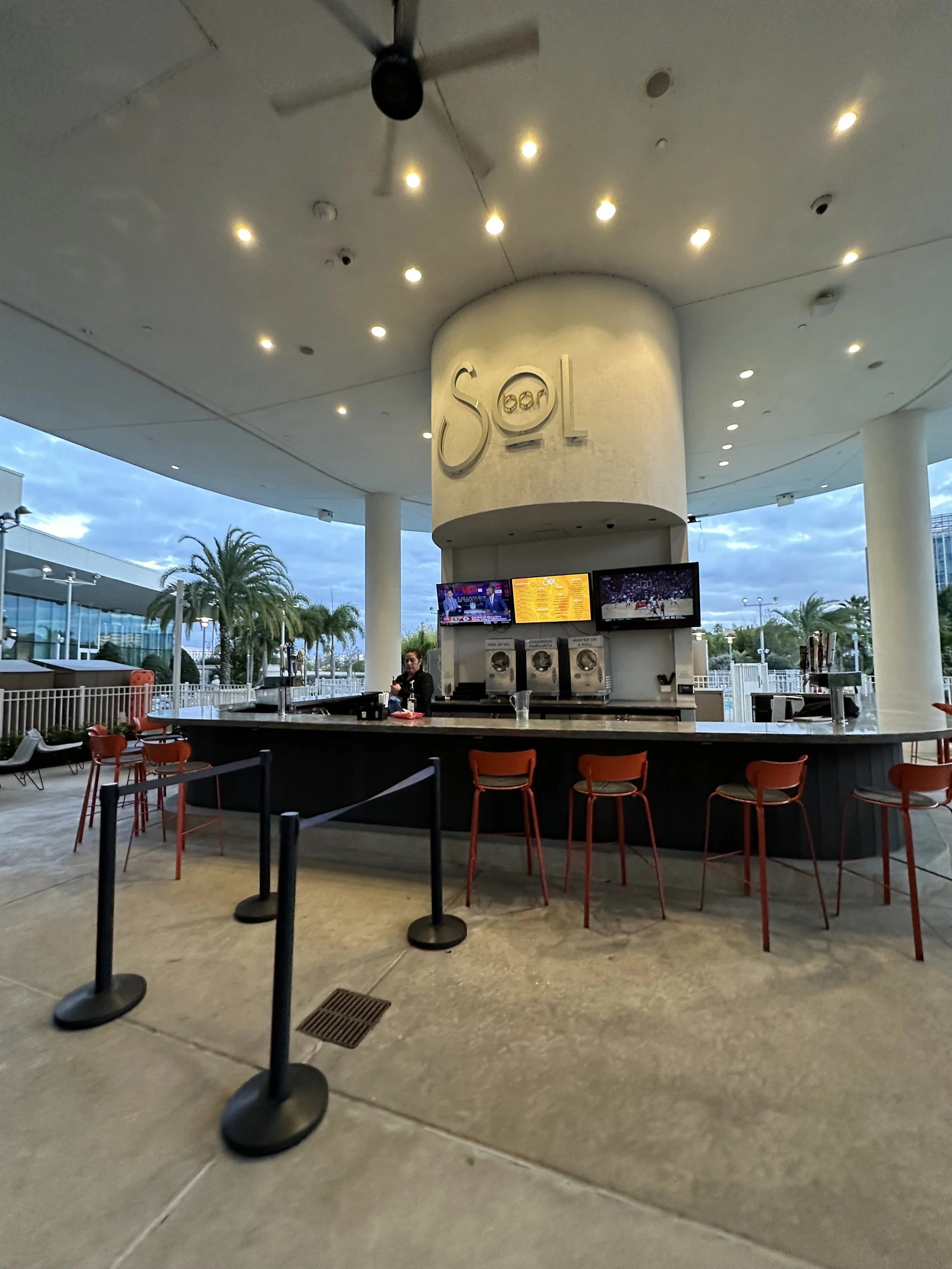 Outdoor bar with red barstools, a bartender, and television screens, featuring the Sol logo on a large cylindrical structure under a roof with ceiling lights, palm trees in the background, and a cloudy sky.