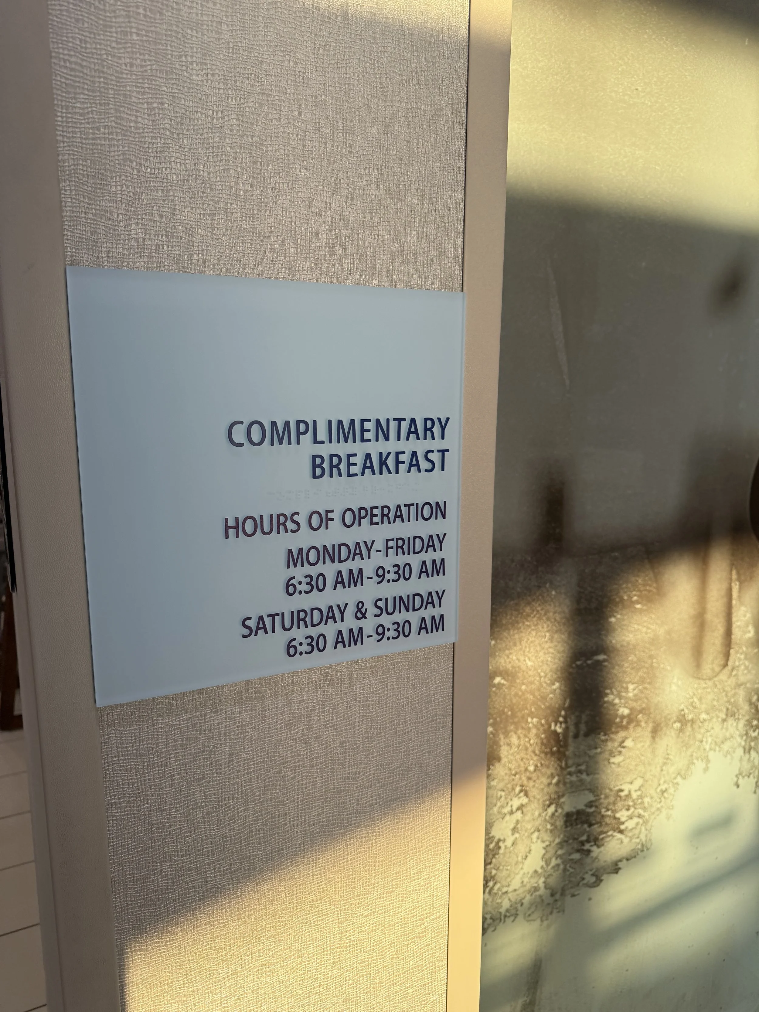 Sign on wall offering complimentary breakfast with hours of operation, Monday to Friday 6:30 AM to 9:30 AM, Saturday and Sunday 6:30 AM to 9:30 AM.