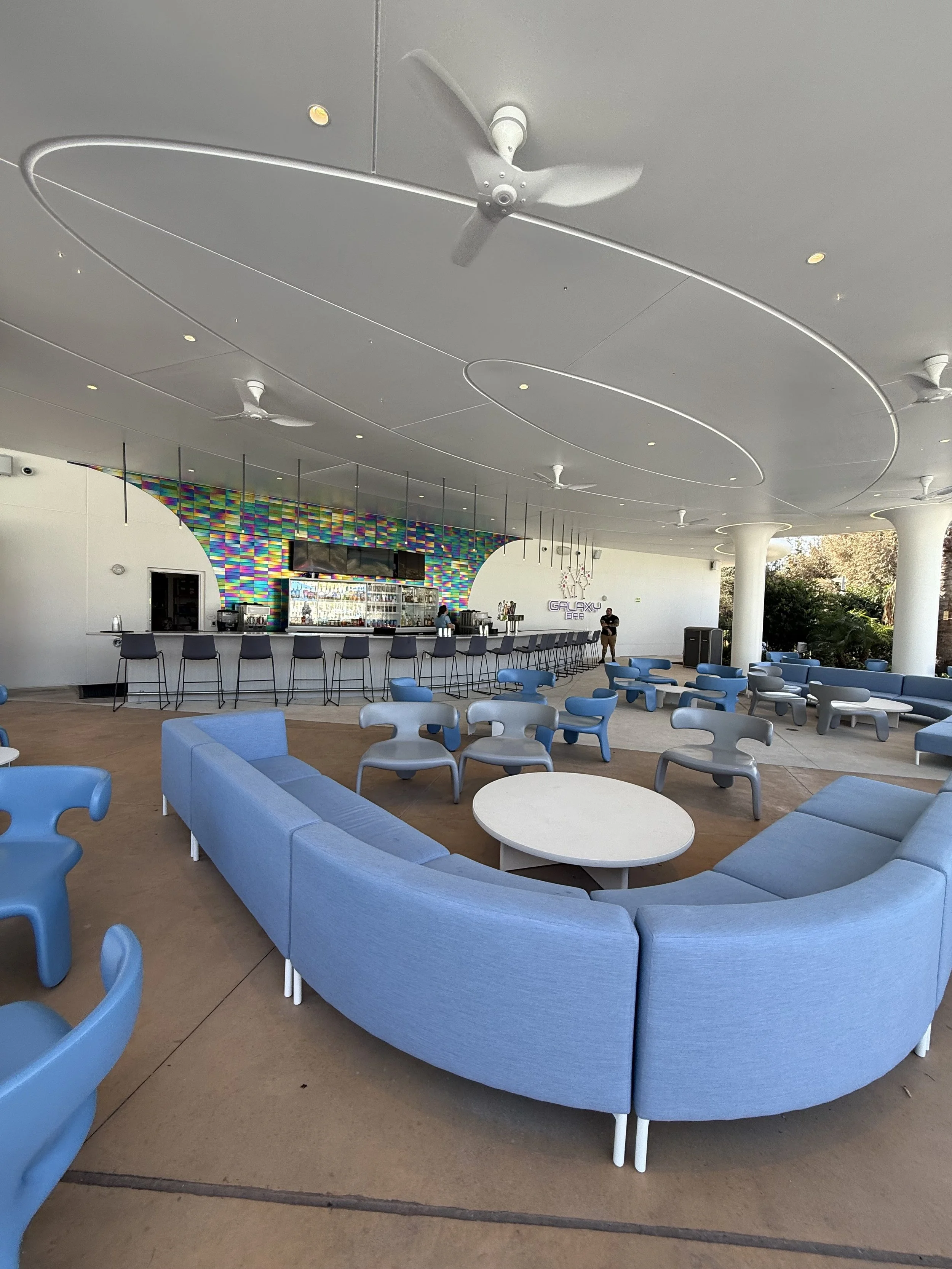 Indoor lounge area with blue curved sofa, multiple small blue and grey chairs, and a bar with holographic wall behind it, under a ceiling with large white ceiling fans.