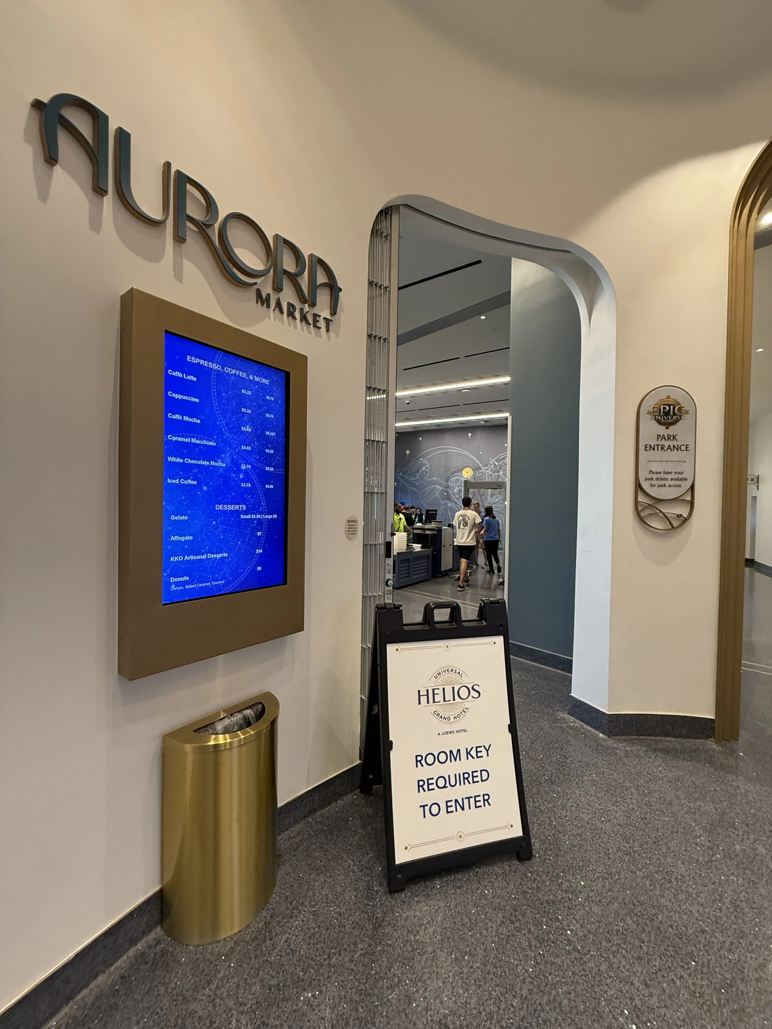 Entrance to the Aura Market in the Universal Helios Grand Hotel. There is a sign that says 'Room Key Required to Enter'. A digital menu board displays coffee and dessert options. There is a gold trash bin beside the menu board.