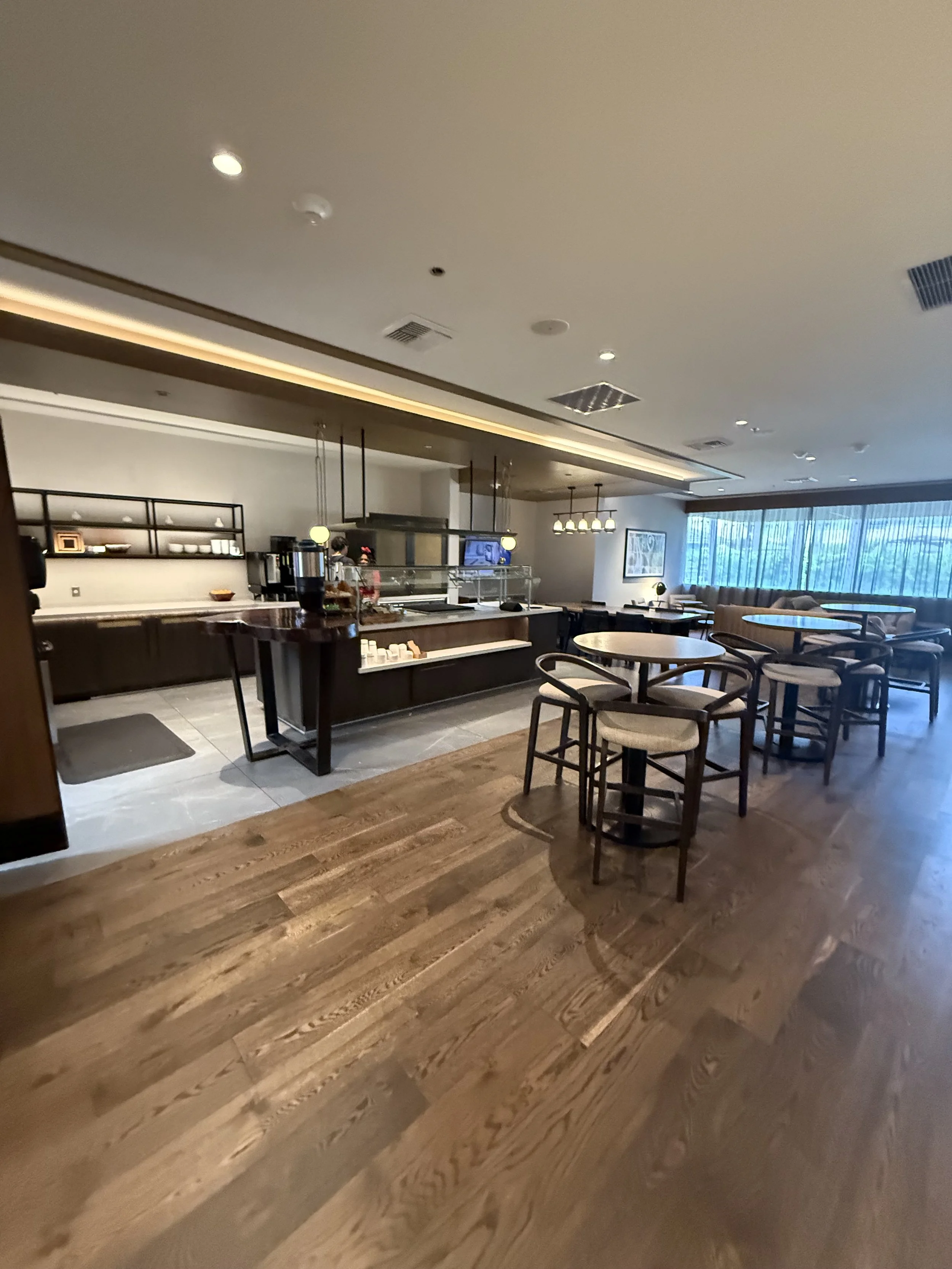 A cozy cafe or lounge area with wooden floors, round tables, and cushioned chairs, near a breakfast or buffet station with shelves, a counter, and a serving area. Large windows with vertical blinds let in natural light.
