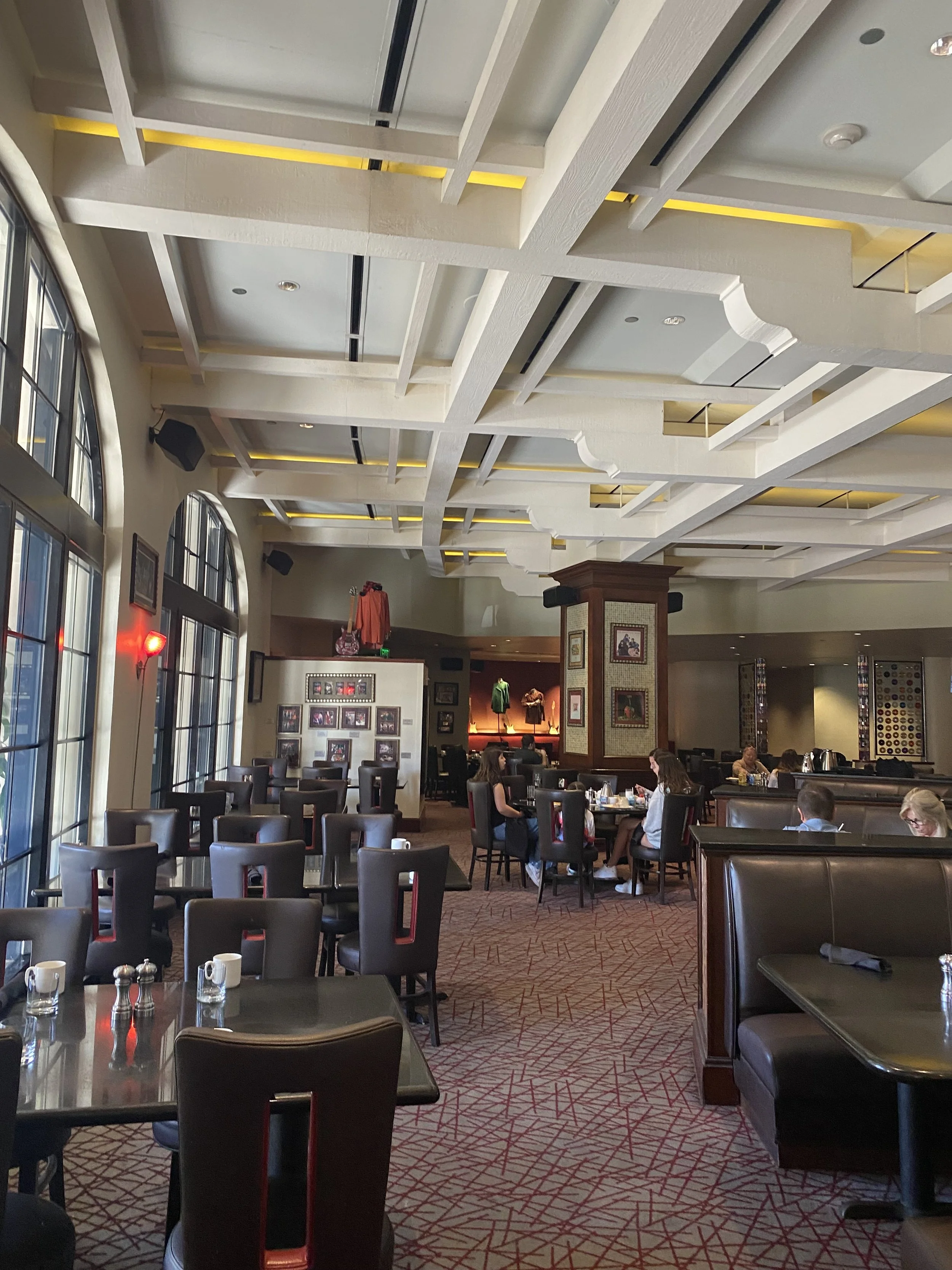 Interior of a restaurant with black chairs, tables set with cups, and a patterned carpet. Large windows on the left, and framed pictures on the walls. Several people are seated and dining.