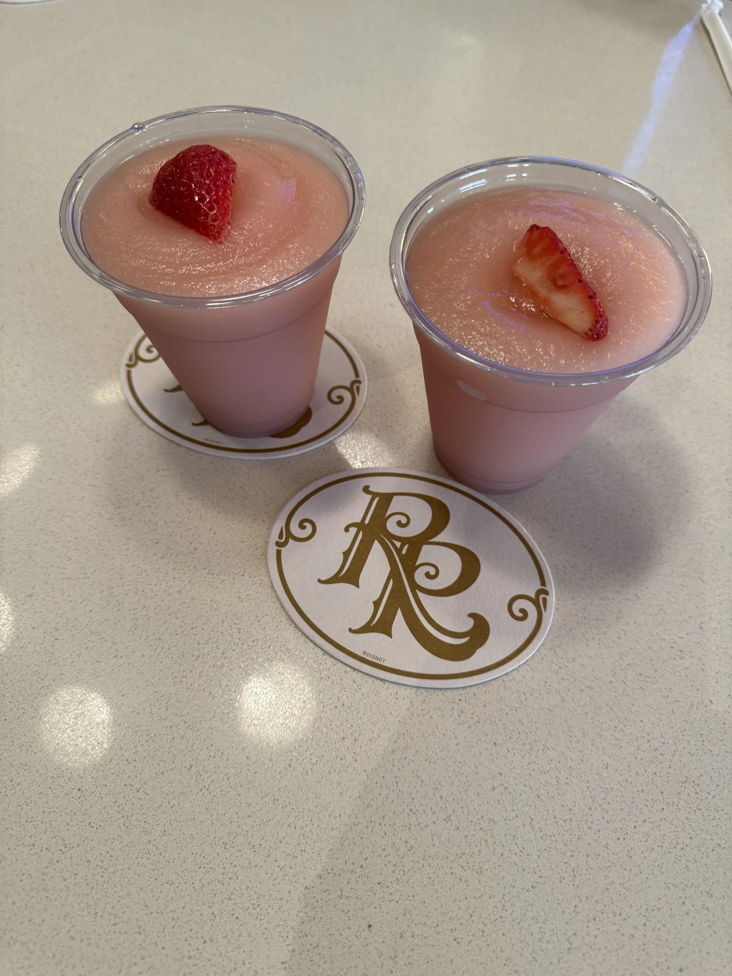 Two pink strawberry smoothies topped with strawberry pieces, placed on round coasters with a gold decorative letter 'R' on a white table.