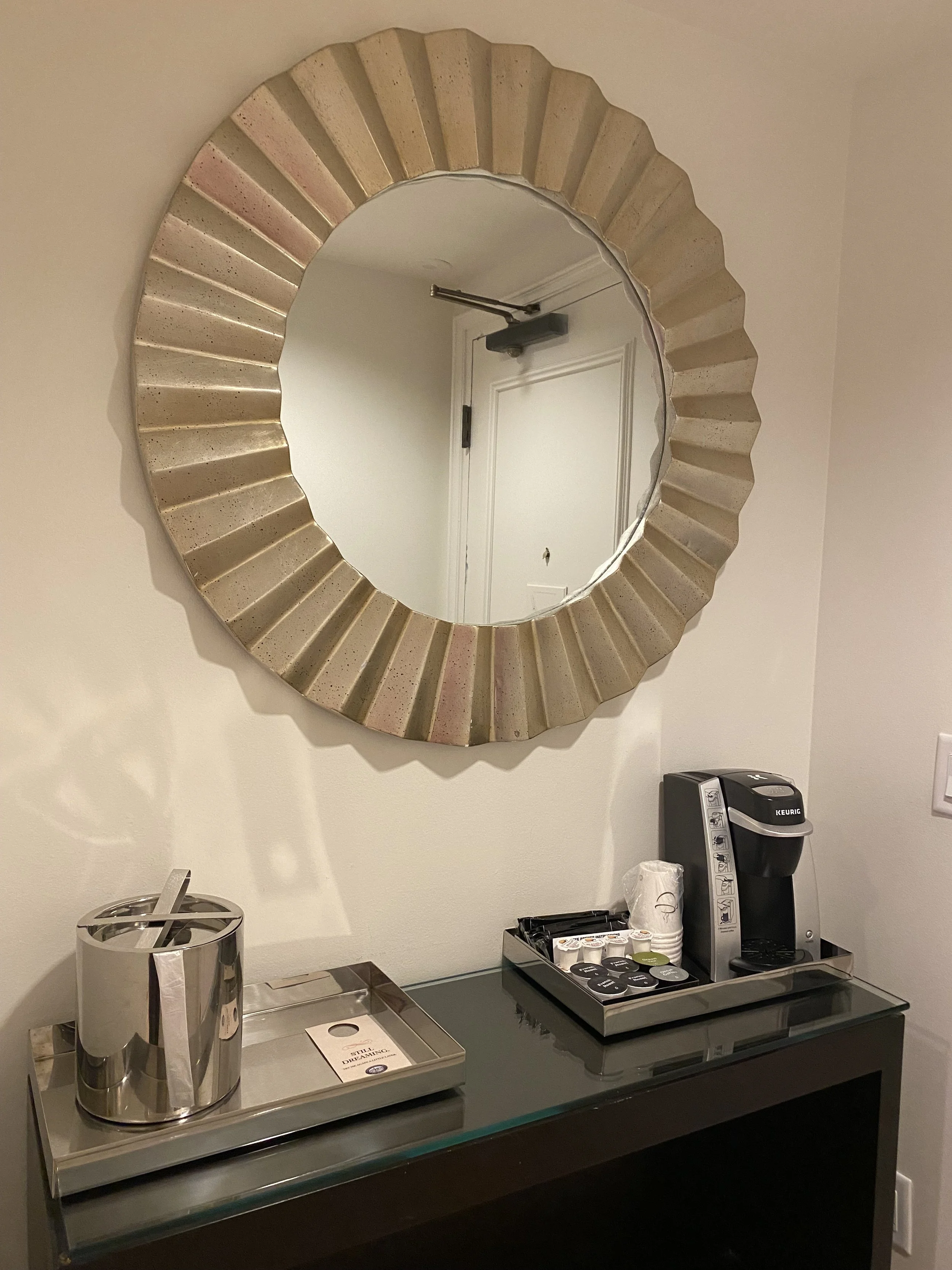 Decorative round mirror with a gold and pinkish geometric frame hanging on a beige wall. Below the mirror is a black console table holding a stainless steel ice bucket, a tray with a paper note, and a Keurig coffee machine on a tray with cups and pod