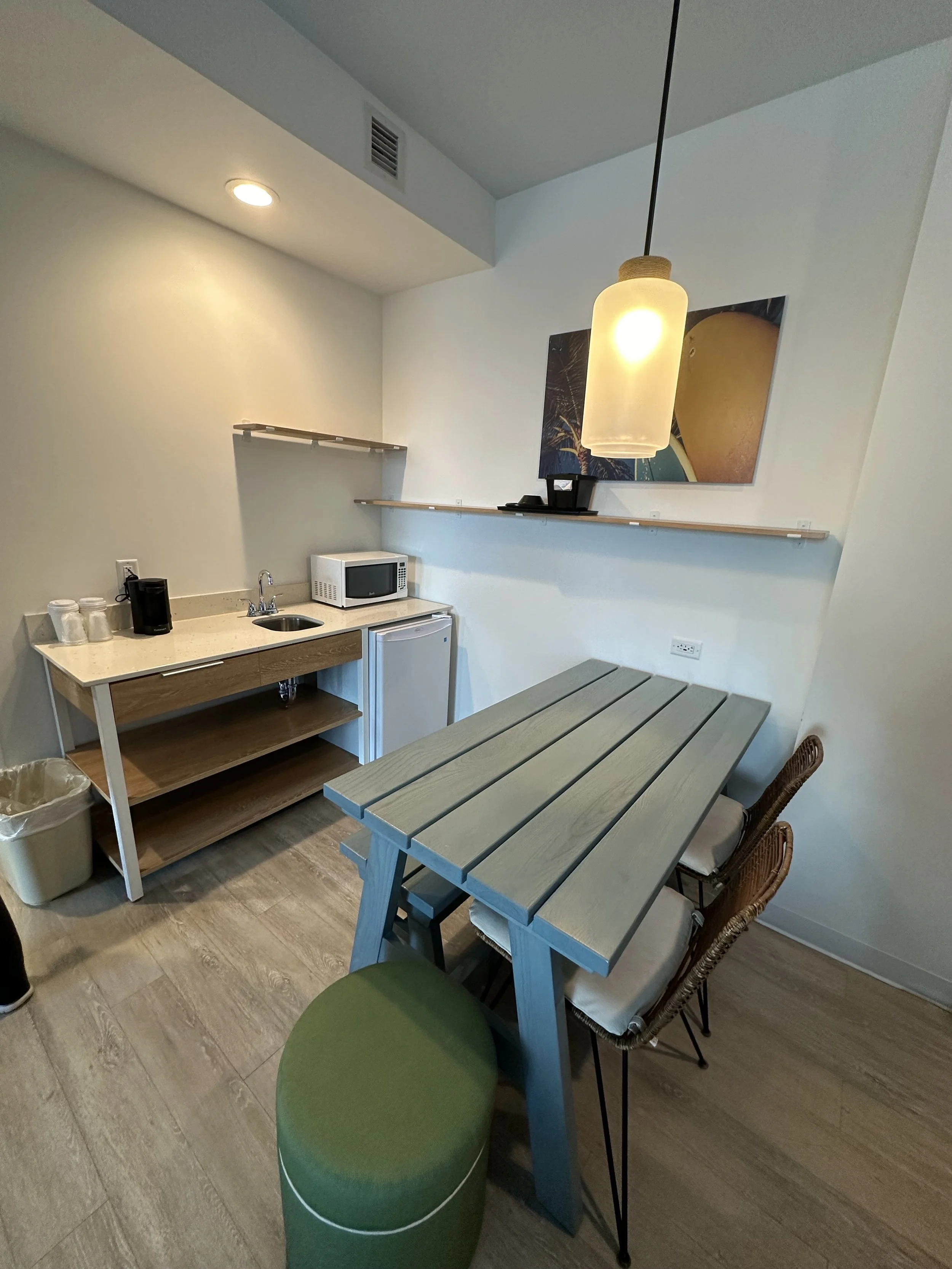 Small kitchenette with a microwave, mini fridge, and a coffee maker, adjacent to a dining area with a wooden table, wicker chairs, and a green stool, with modern artwork and a hanging pendant light.