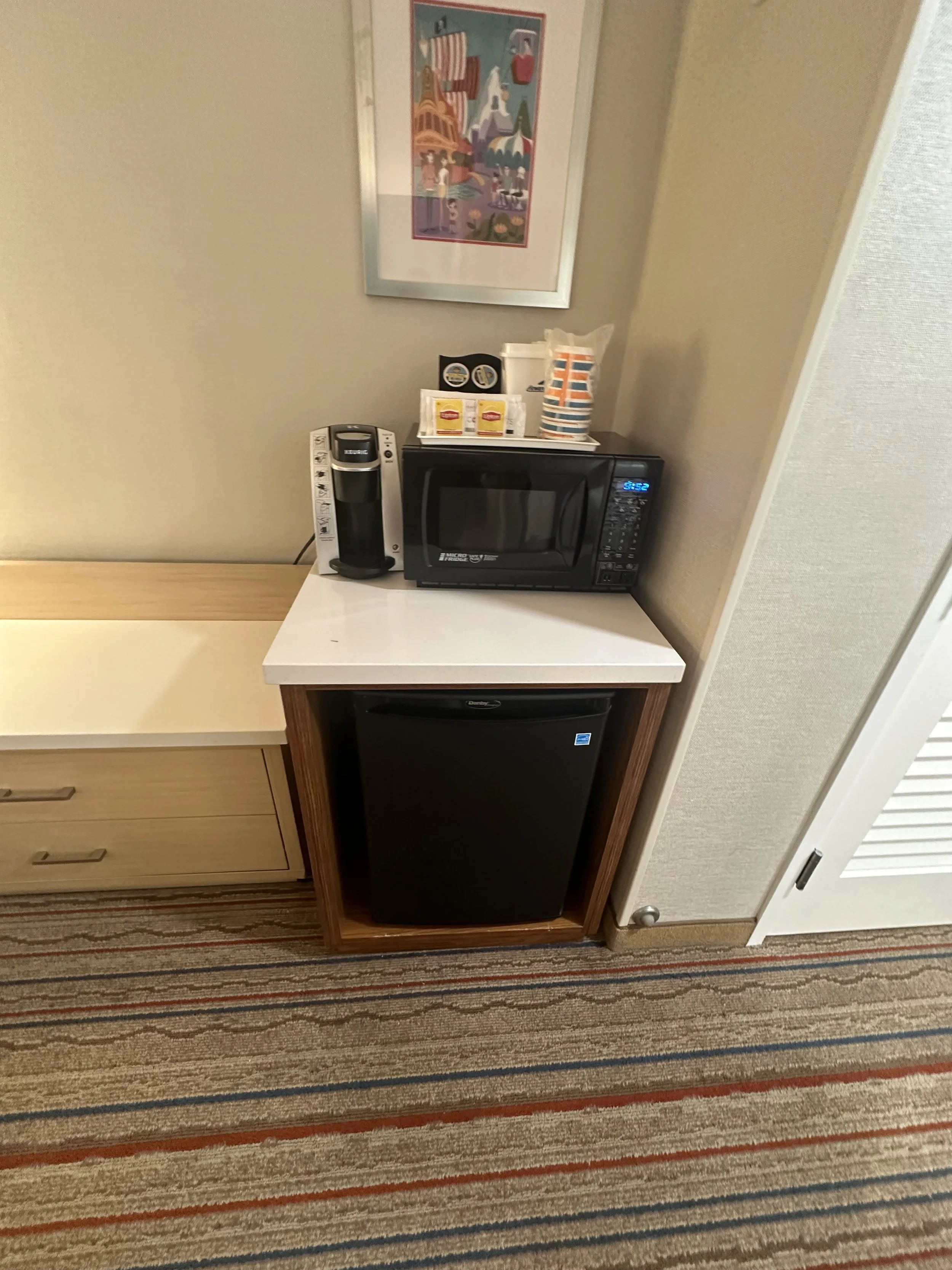 A hotel room kitchenette with a small black mini fridge beneath a white counter, a microwave oven, a coffee maker, and a tray with coffee supplies on top of the microwave. There is a framed colorful poster on the wall above the counter.