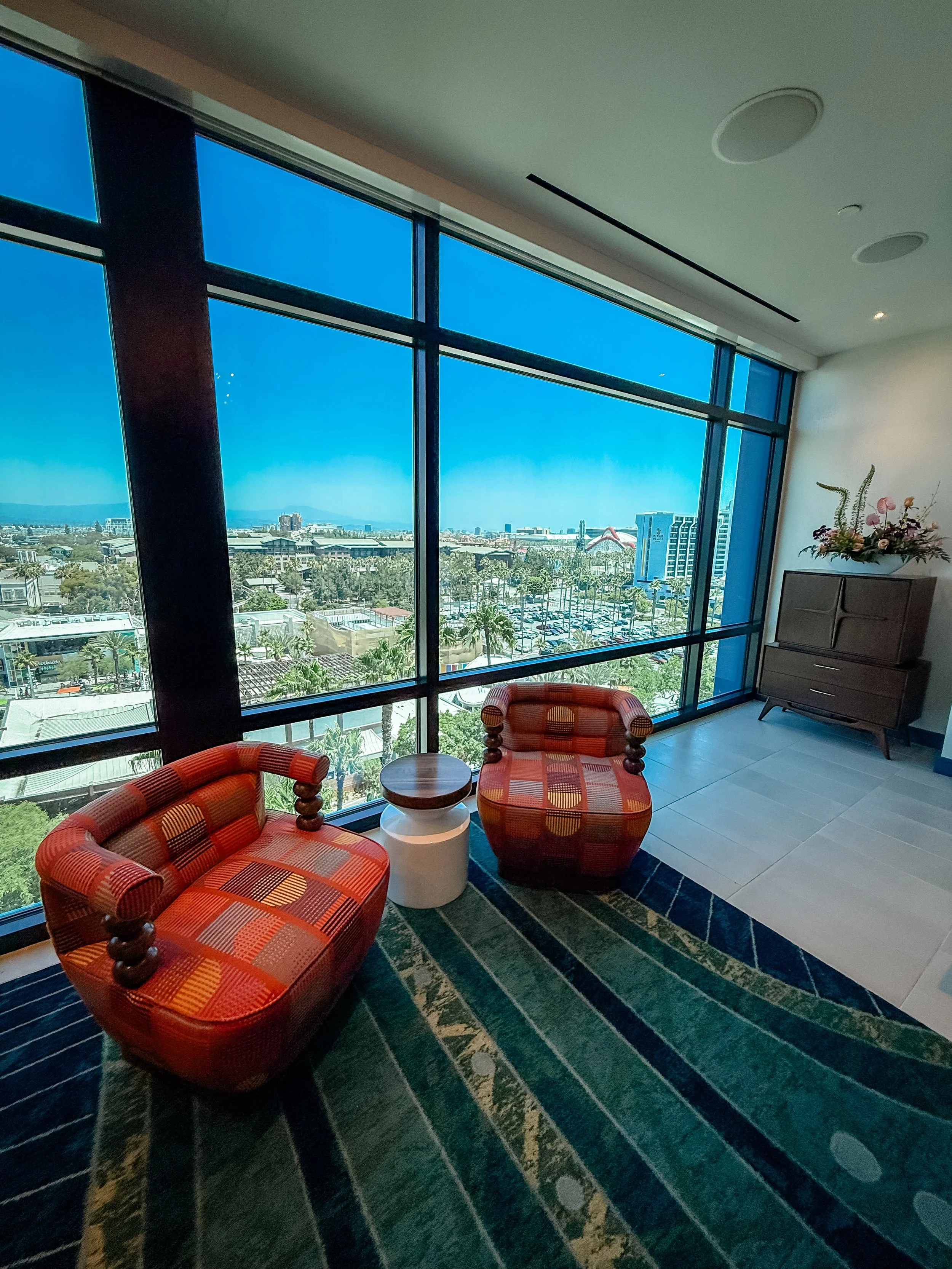 Two red patterned chairs with rounded arms and wooden accents sit next to a small round wooden table in front of a large window with a view of a cityscape, parking lot, and blue sky.