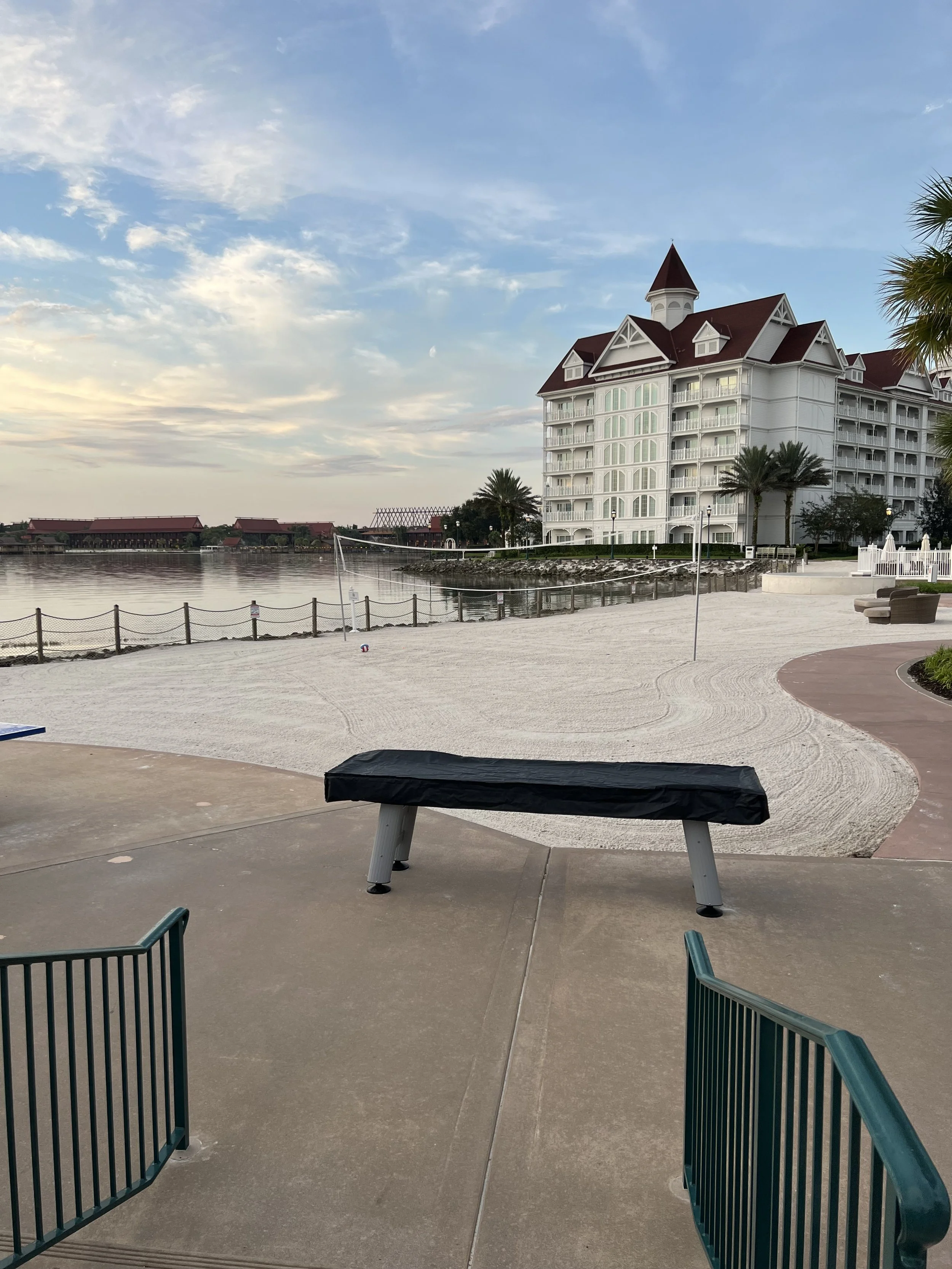 View of a sandy beach area near a waterfront with a large white multi-story hotel or resort building in the background, palm trees, and a partly cloudy sky during either sunrise or sunset.