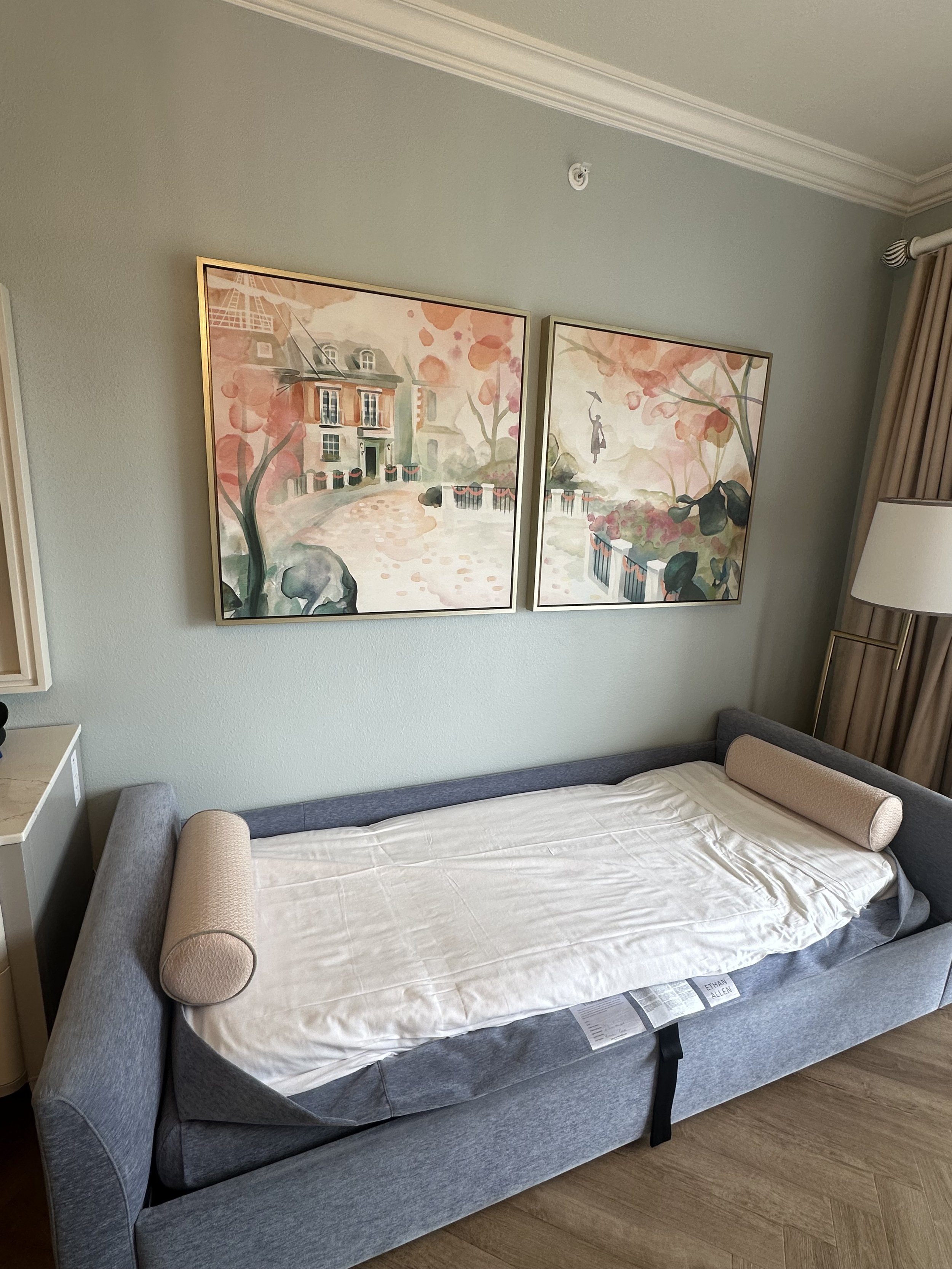 A hotel room with a sofa bed, decorative pillows, artwork on the wall, a nightstand, and curtains.