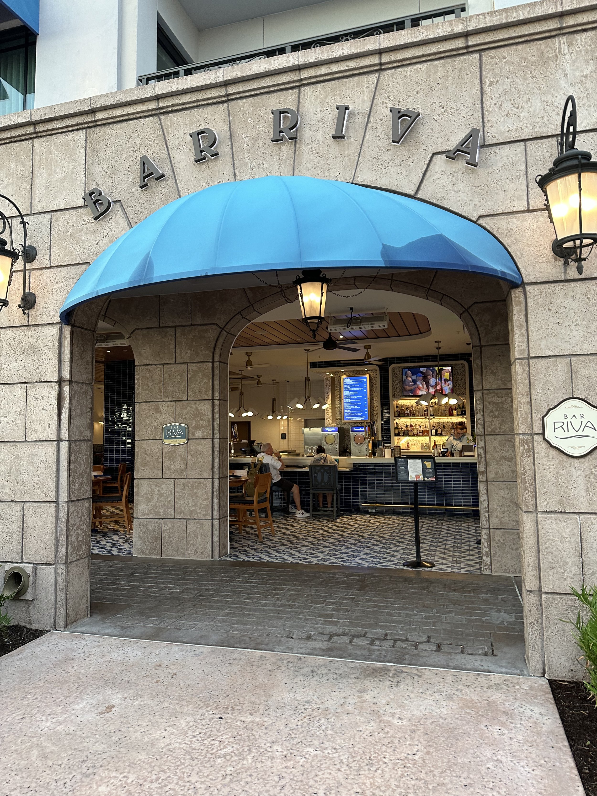 Entrance to a restaurant called Bar Riva with a blue awning and illuminated lamps on either side. Inside, there are bar stools, a digital menu screen, and a bar counter with bottles on display.