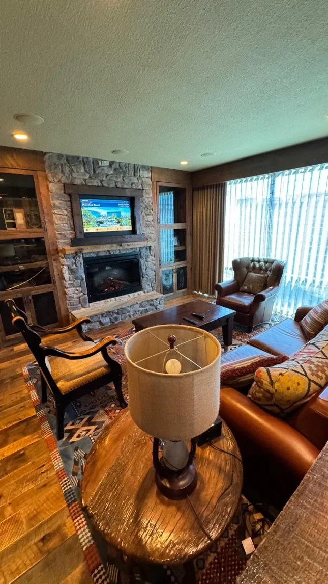 Living room with stone fireplace, wall-mounted TV, large window with vertical blinds, leather armchair, patterned sofa, wooden coffee table, round side table with lamp, and wooden floors.