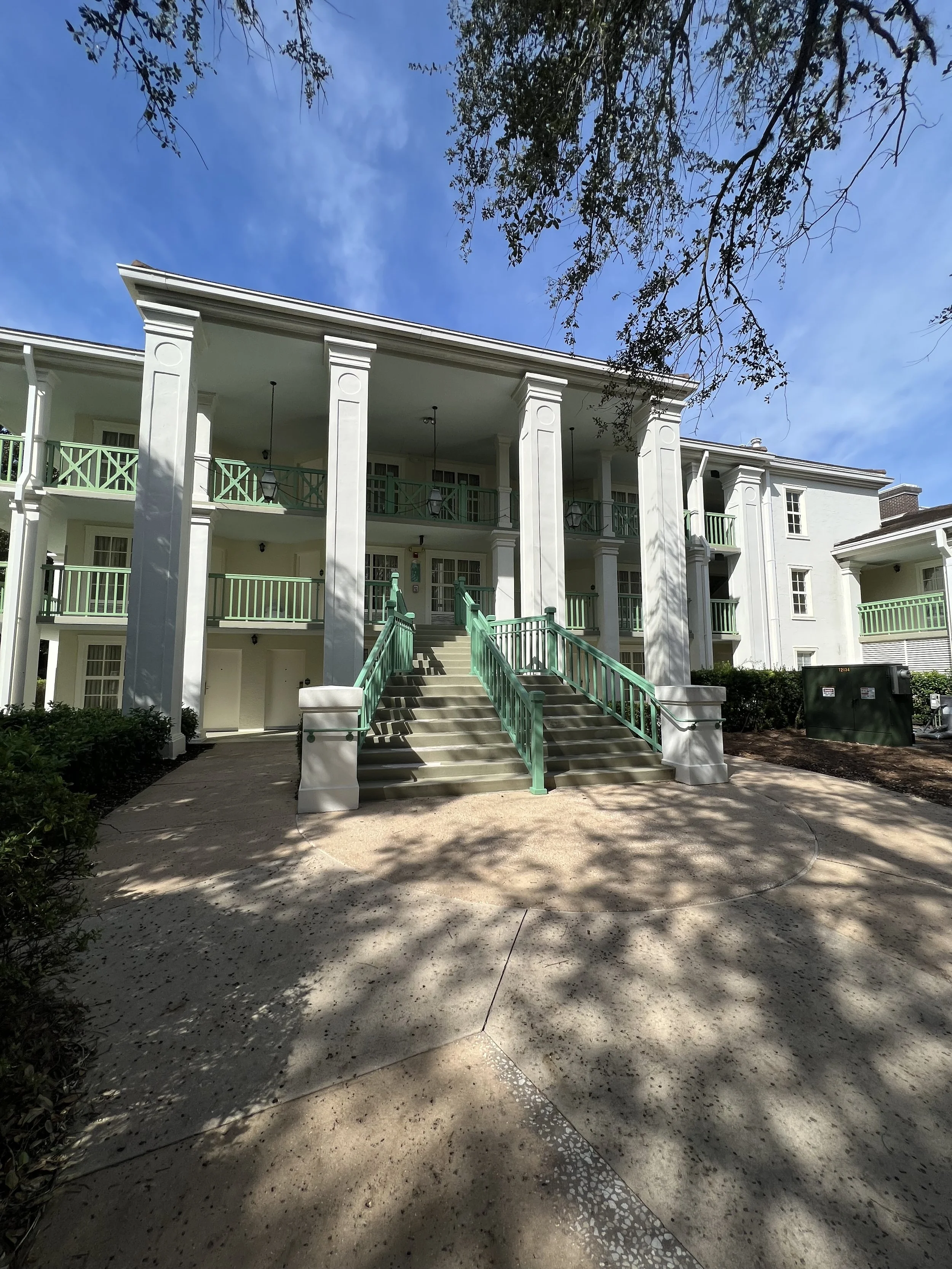 Exterior view of a white apartment building with green railings and a staircase leading to the main entrance, under a blue sky with some tree shadows.