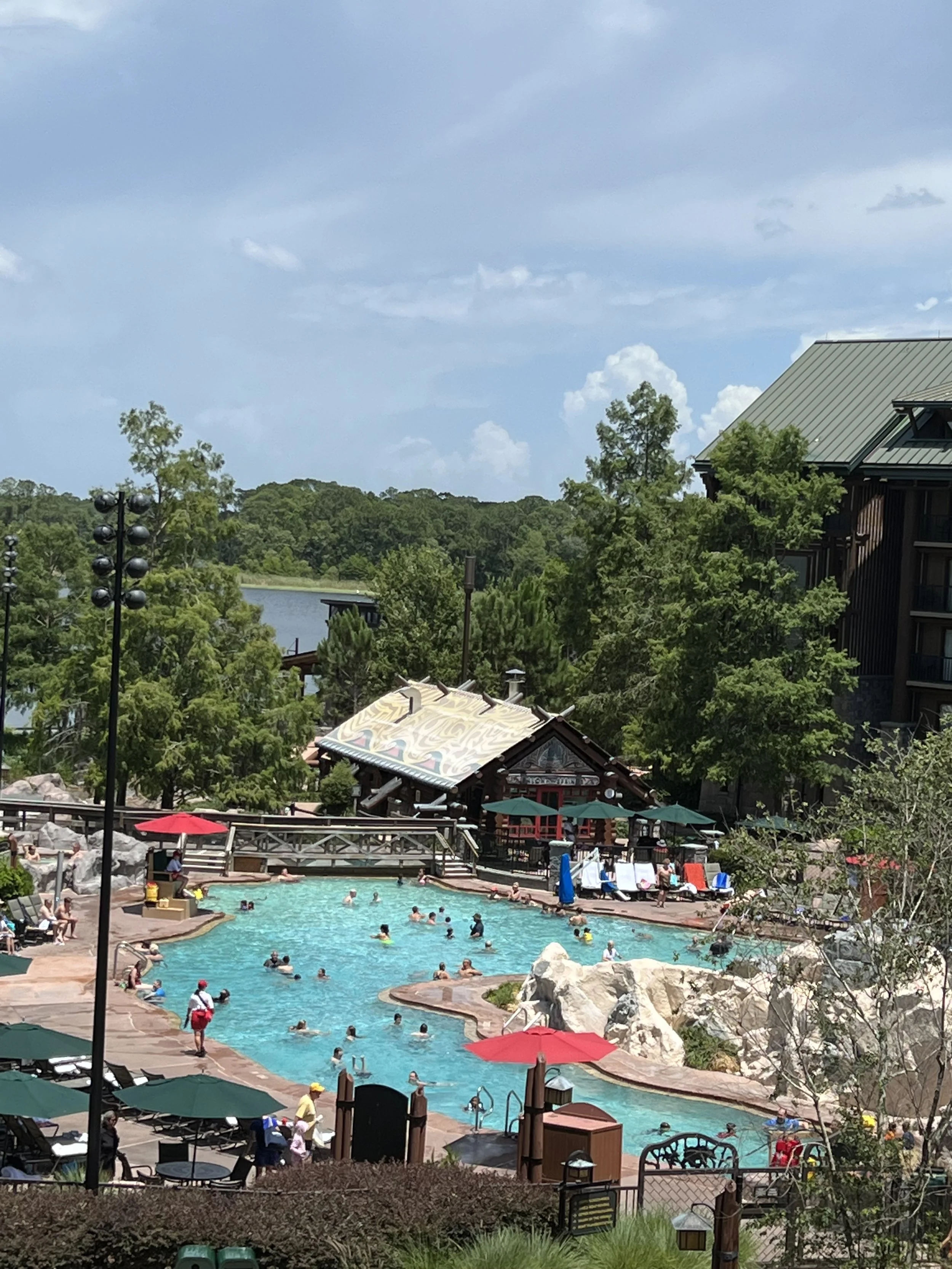 People swimming and lounging by a large outdoor swimming pool with umbrellas and trees, a small building, and a lake in the background on a sunny day.
