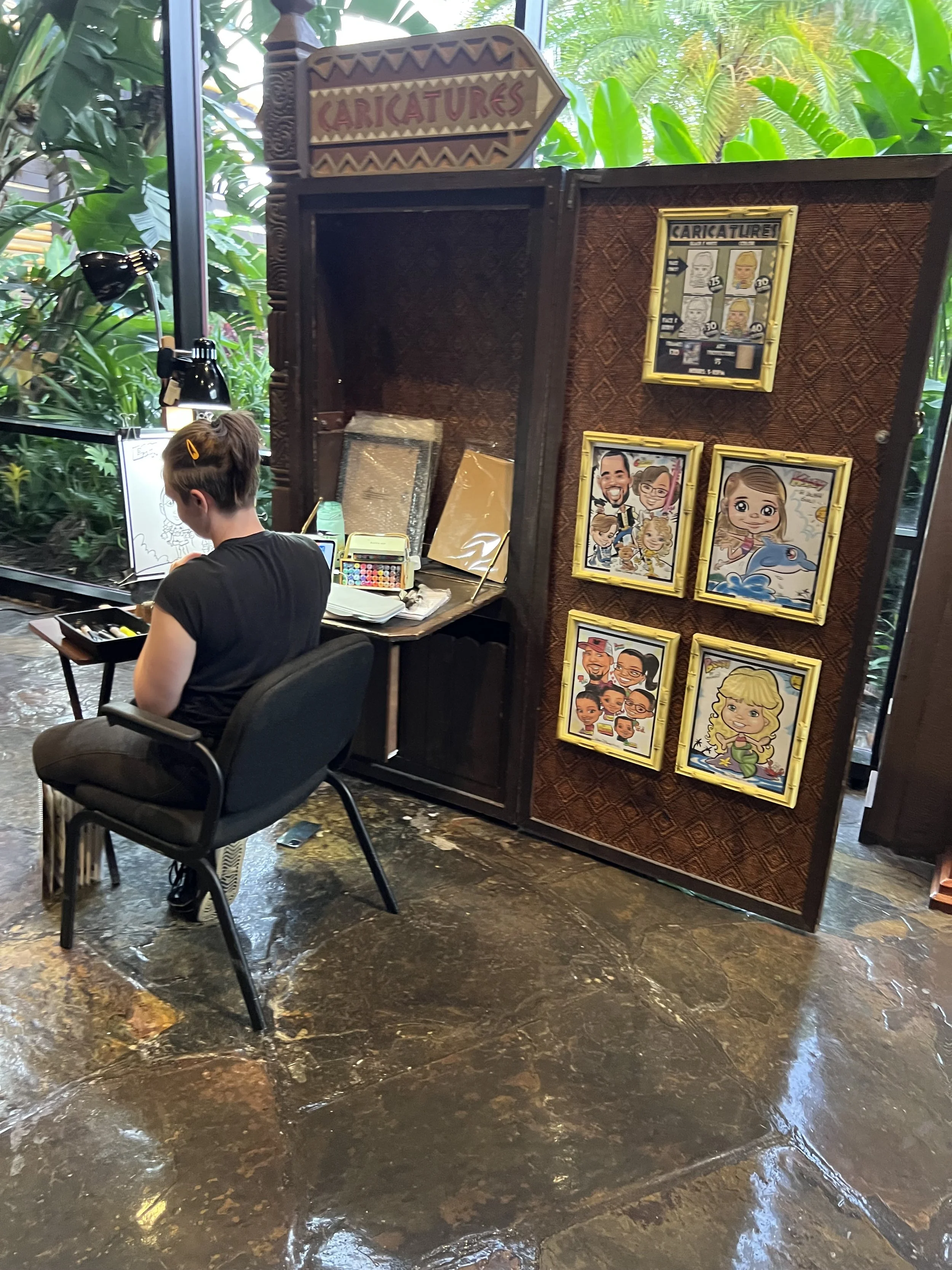 A woman sitting at a desk creating caricature drawings with art supplies, in a tropical-themed indoor space with green plants outside the window. A display board showcases framed caricature portraits and a sign indicating 'Caricatures'.