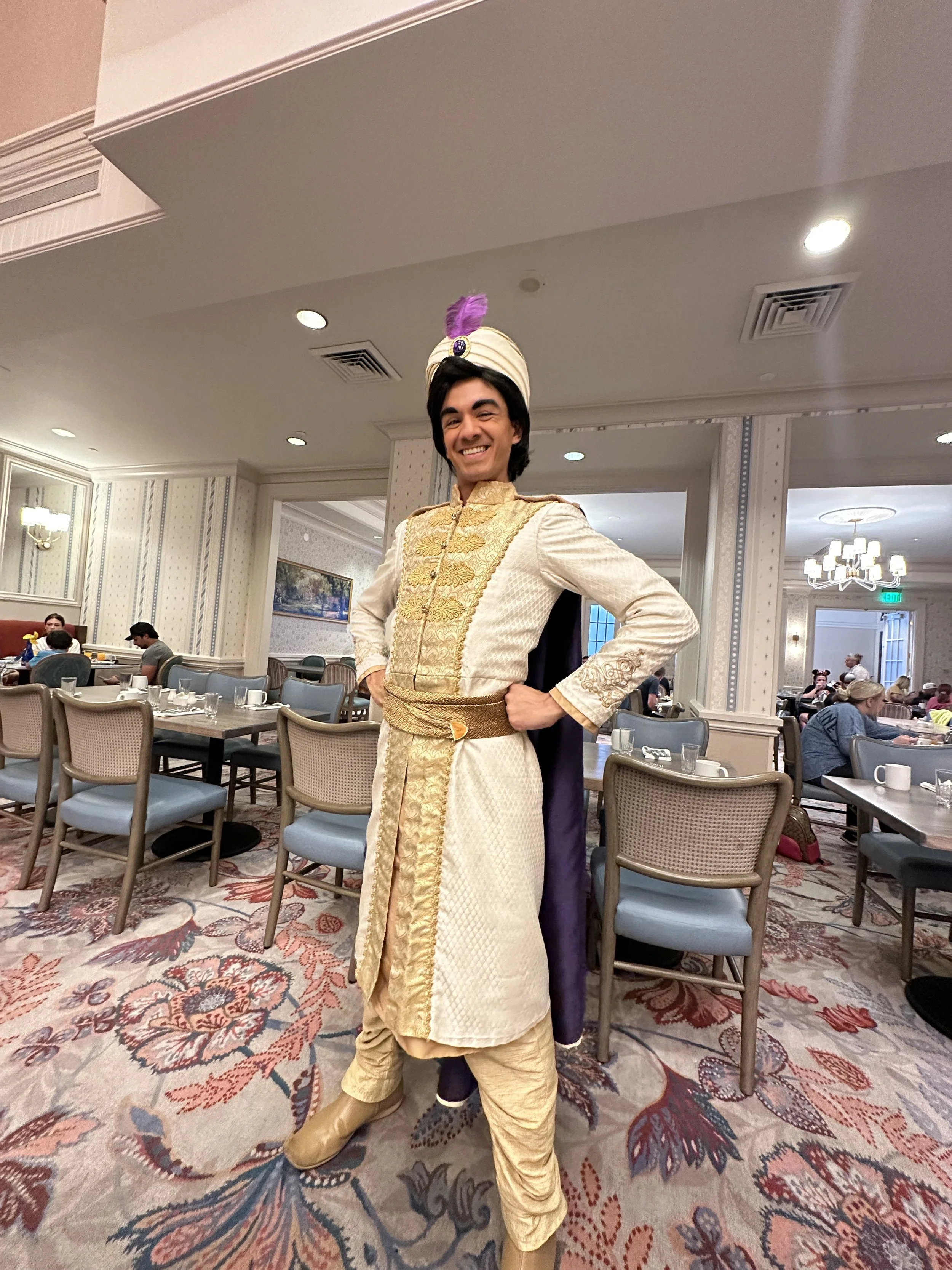 A man dressed in an ornate, traditional Middle Eastern costume, including a cream-colored garment with gold embroidery, a matching turban with a purple feather, and a dark cape, standing confidently in a restaurant dining area.
