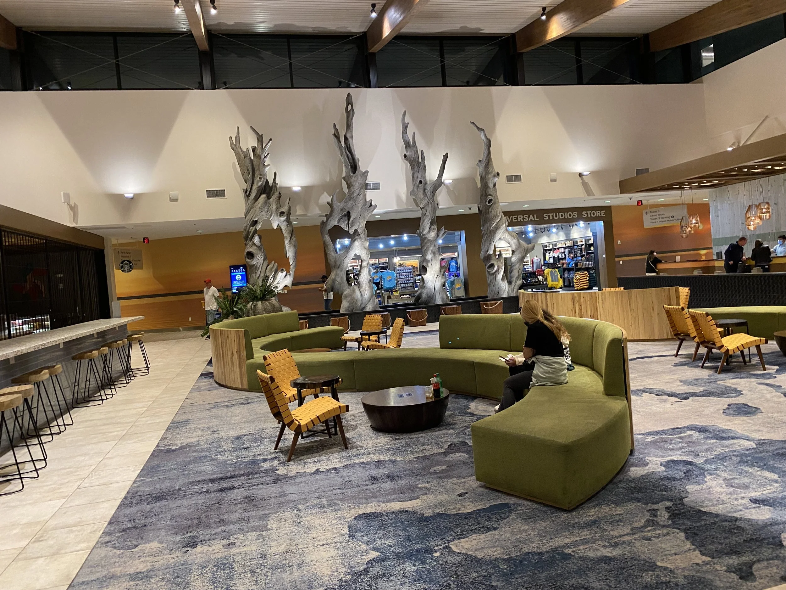 A spacious indoor lounge area with green curved couches, small wooden chairs with yellow and orange upholstery, and a woman sitting on the couch using her phone. In the background, there are large artistic tree sculptures and a Universal Studios stor