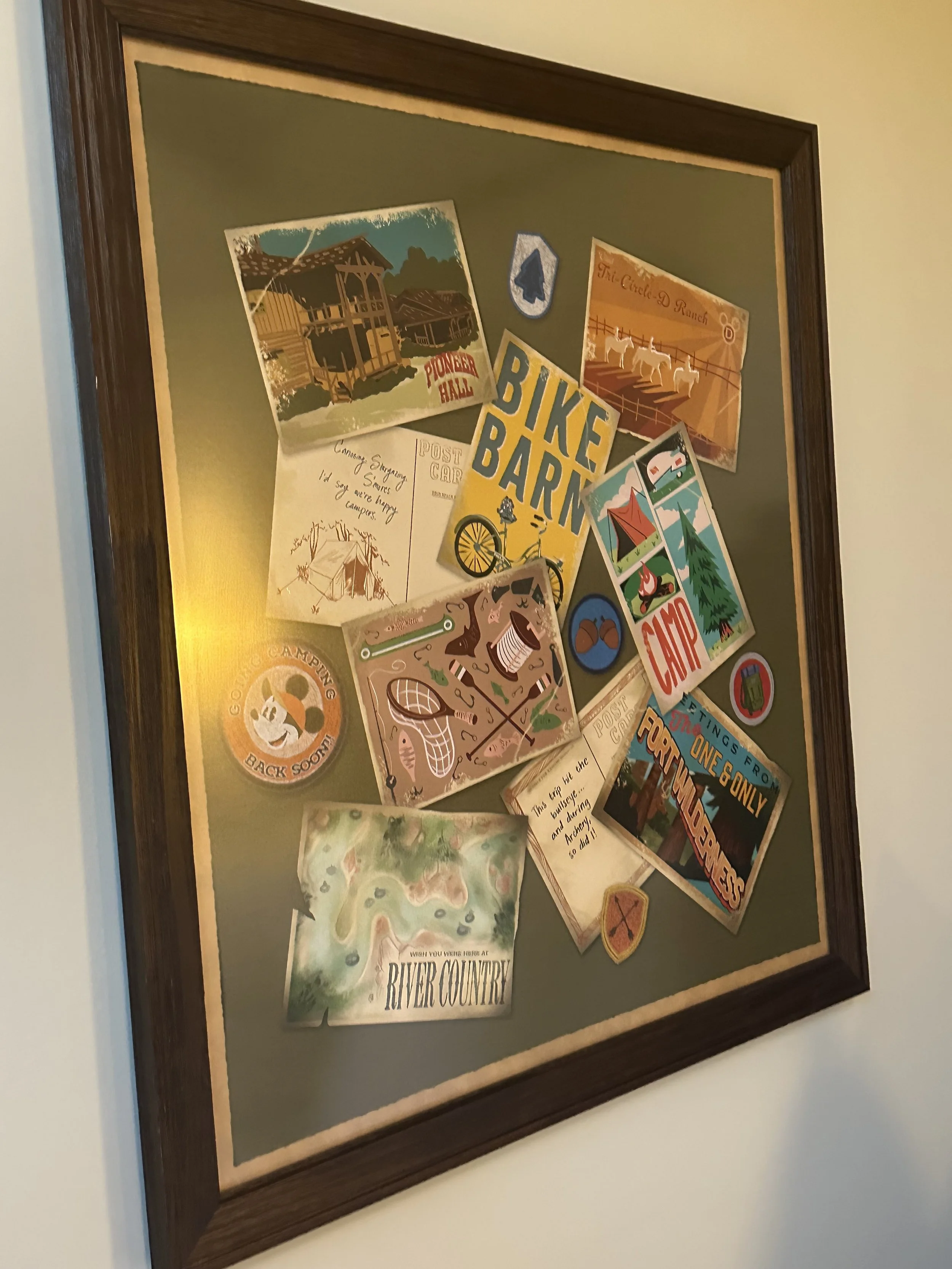 Collection of camping and outdoor-themed stickers and postcards on a green background framed on the wall.