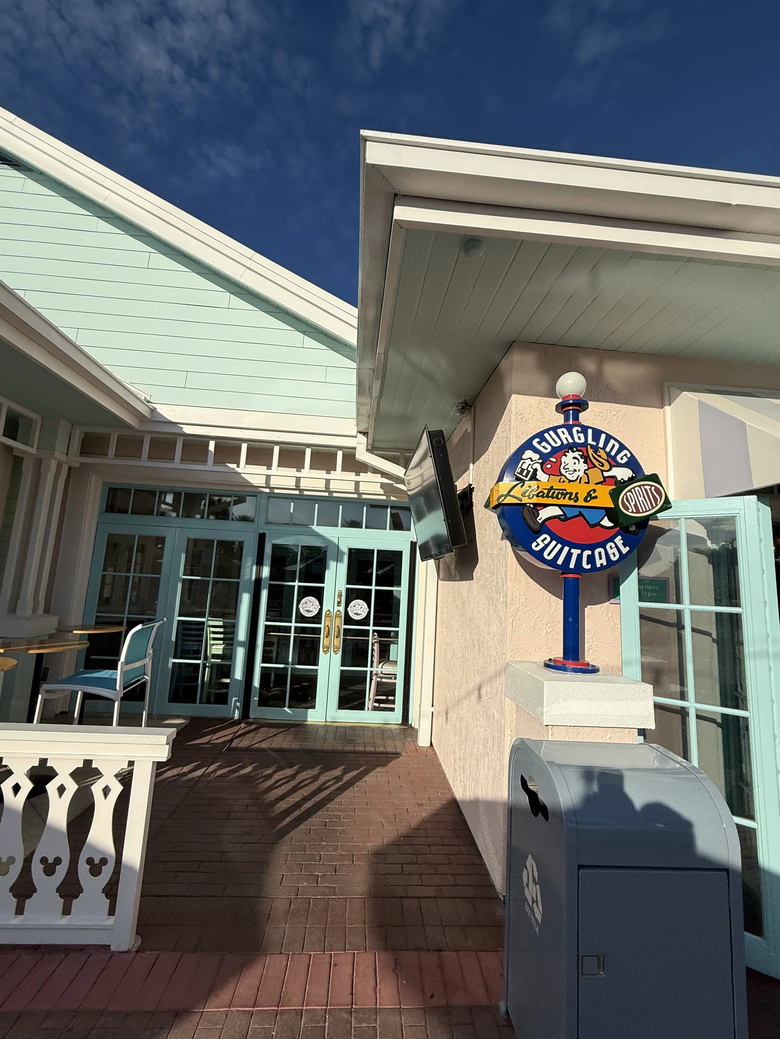 Entrance to a storefront with a sign that reads 'Gurgling Suitcase Liquors & Spirits'. There is a shadow cast on the brick walkway in front of the door, and a recycling bin is visible near the wall.