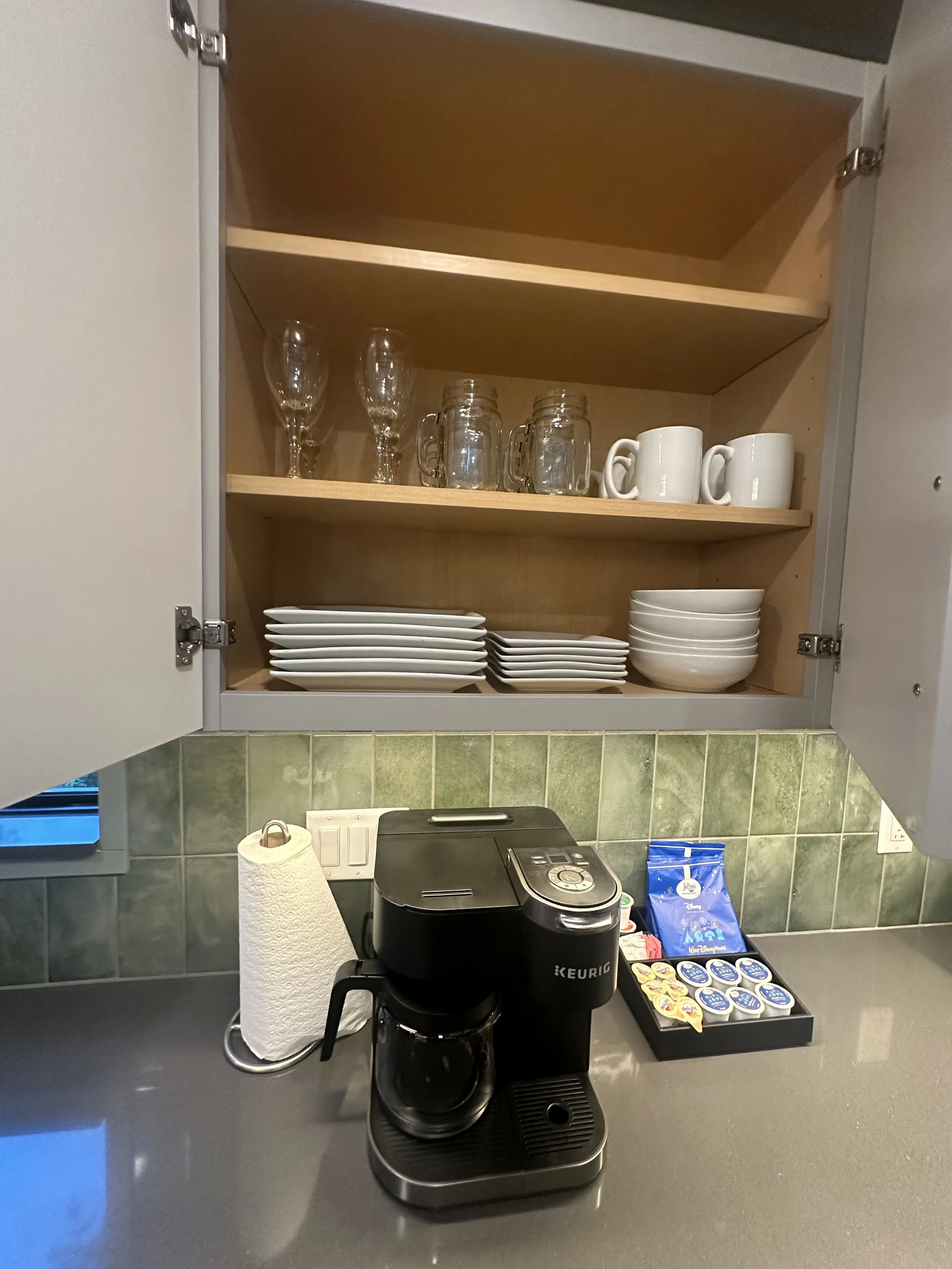 Open kitchen cabinet with glassware, mugs, and dishes; coffee maker and supplies on countertop