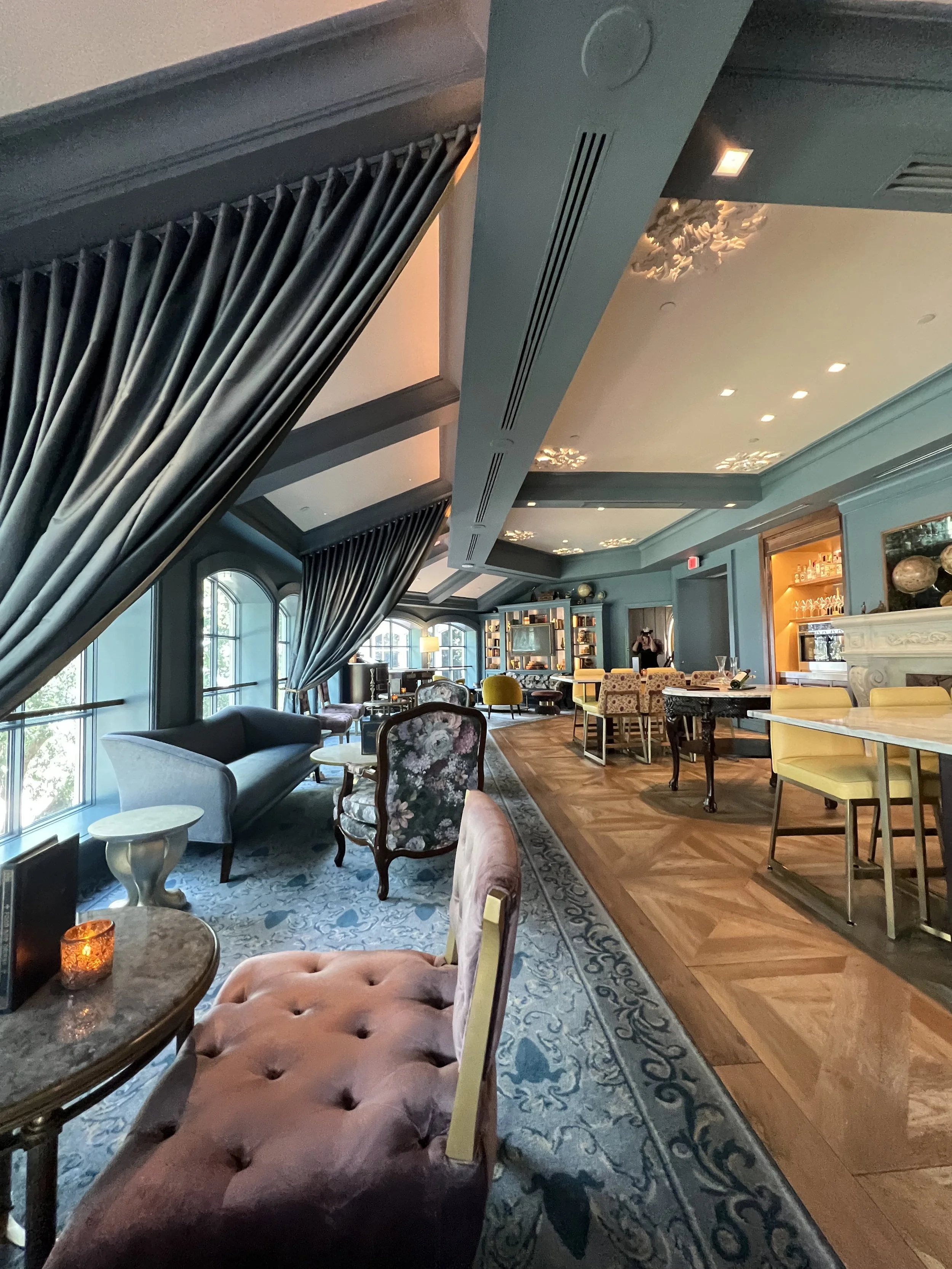 Elegant lounge area with large windows, blue walls, various antique and modern furniture, and decorative ceiling accents.