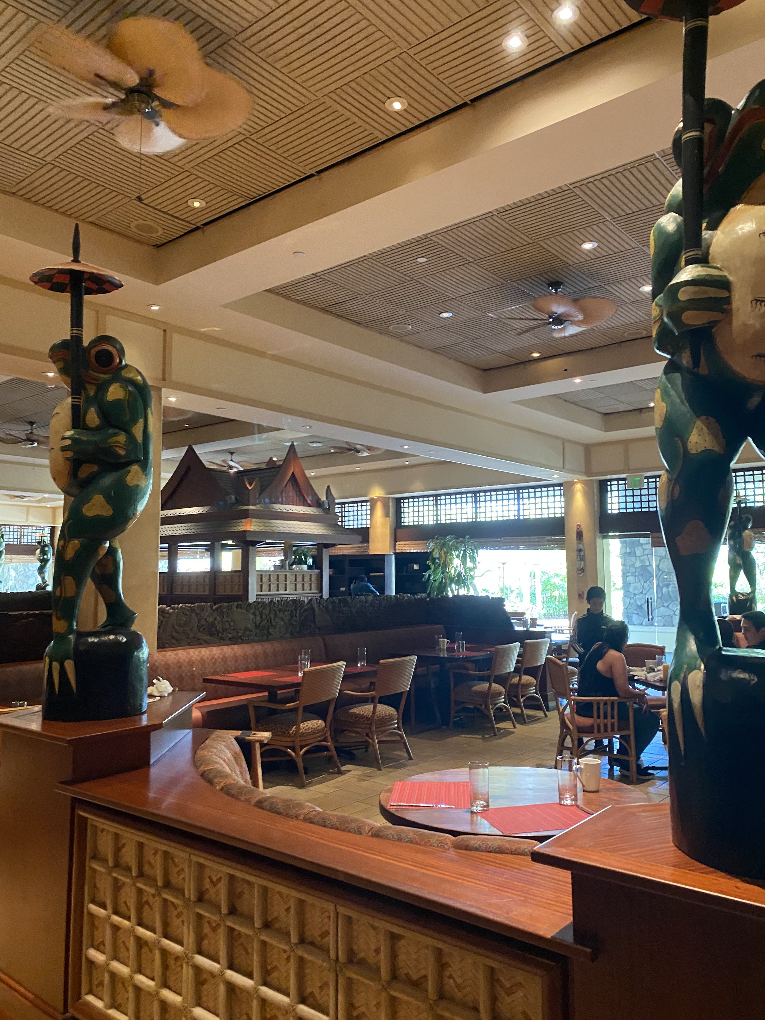 Interior of a restaurant with wooden decor, carved animal statues holding umbrellas, tables set with red placemats and glasses, and a few patrons dining.