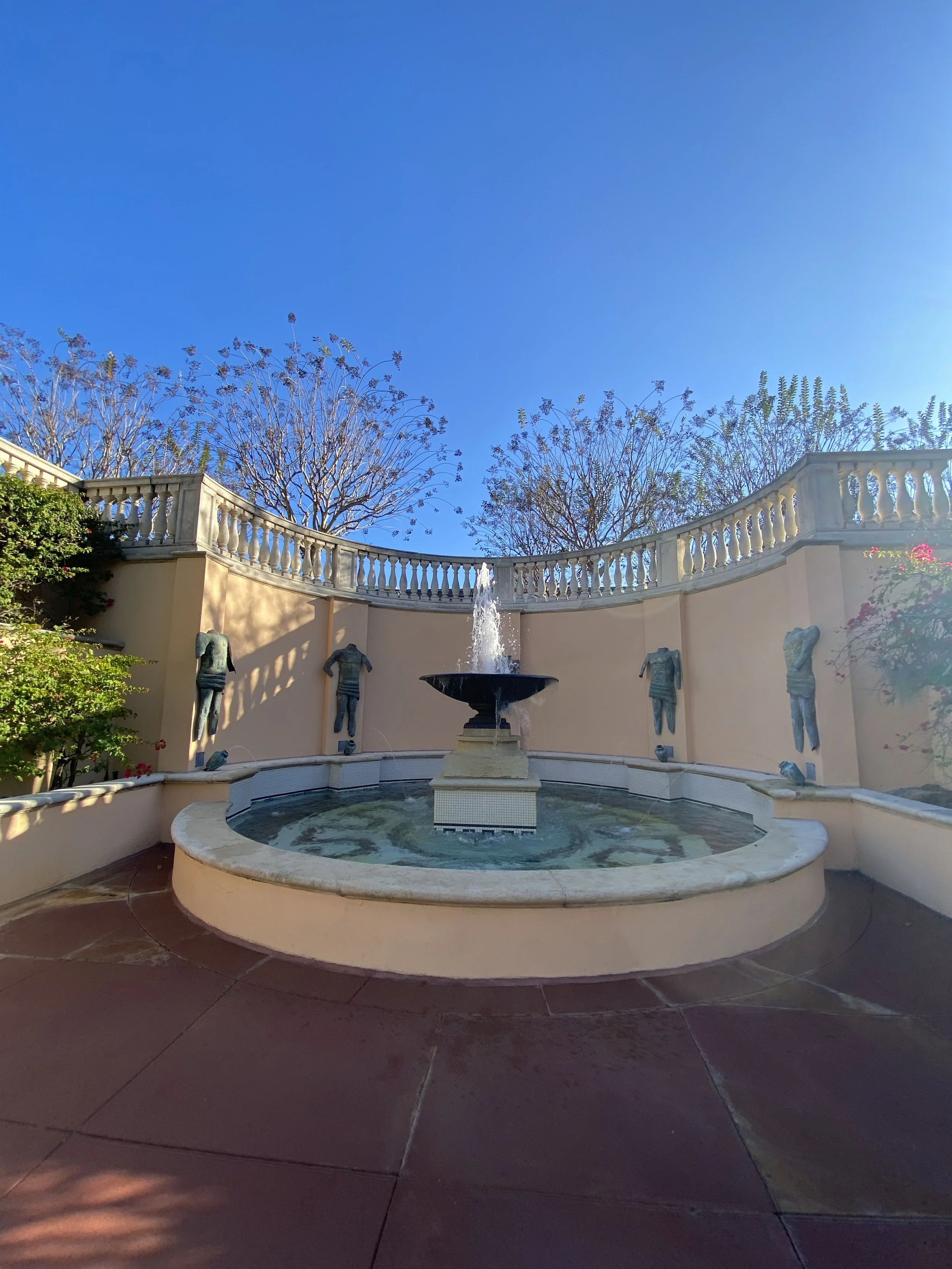 Photo of a fountain in a courtyard with a curved beige wall, decorative sculptures, and a clear blue sky.
