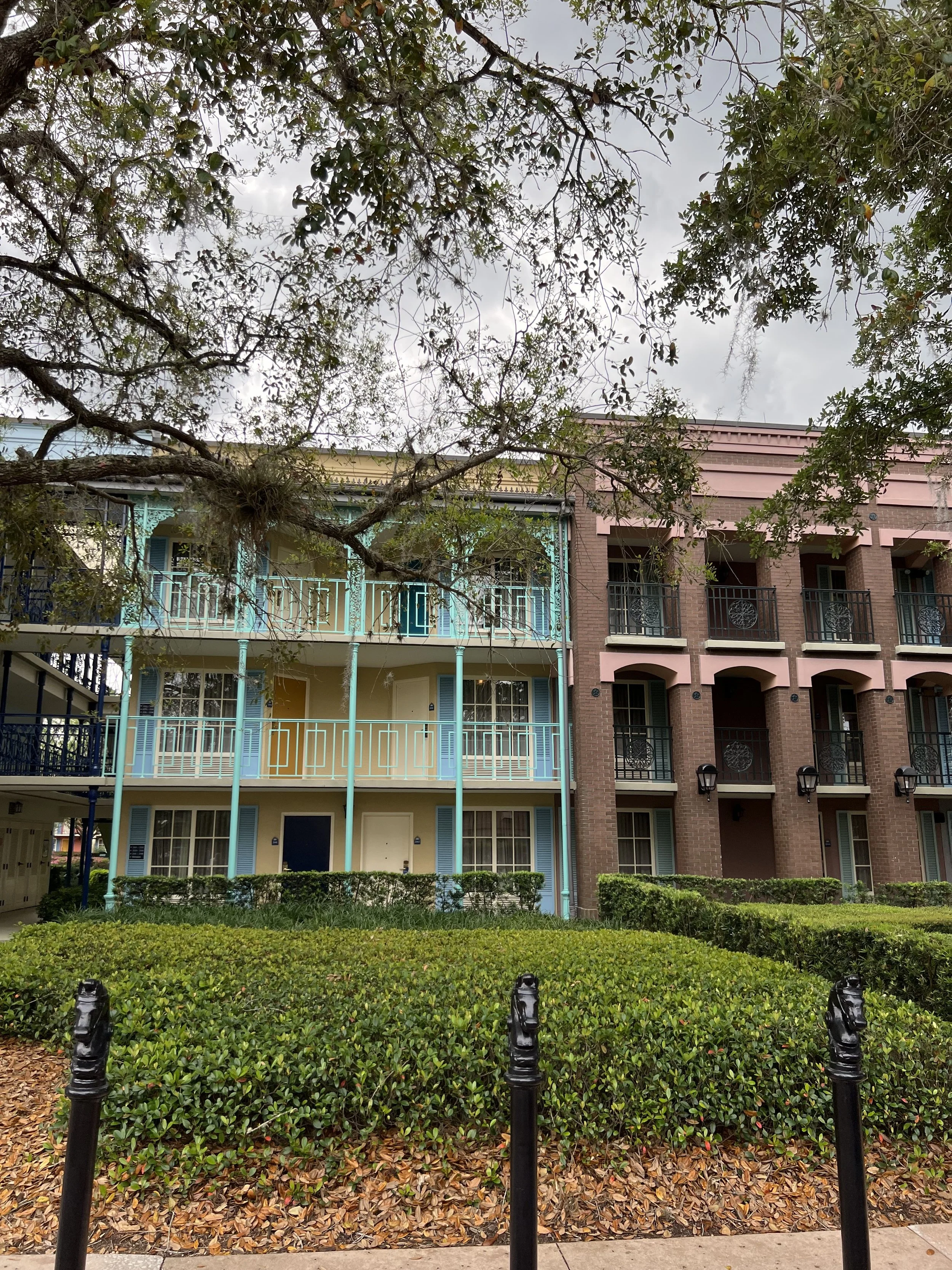 Colorful apartment buildings with balconies, surrounded by green bushes and trees, under a cloudy sky.