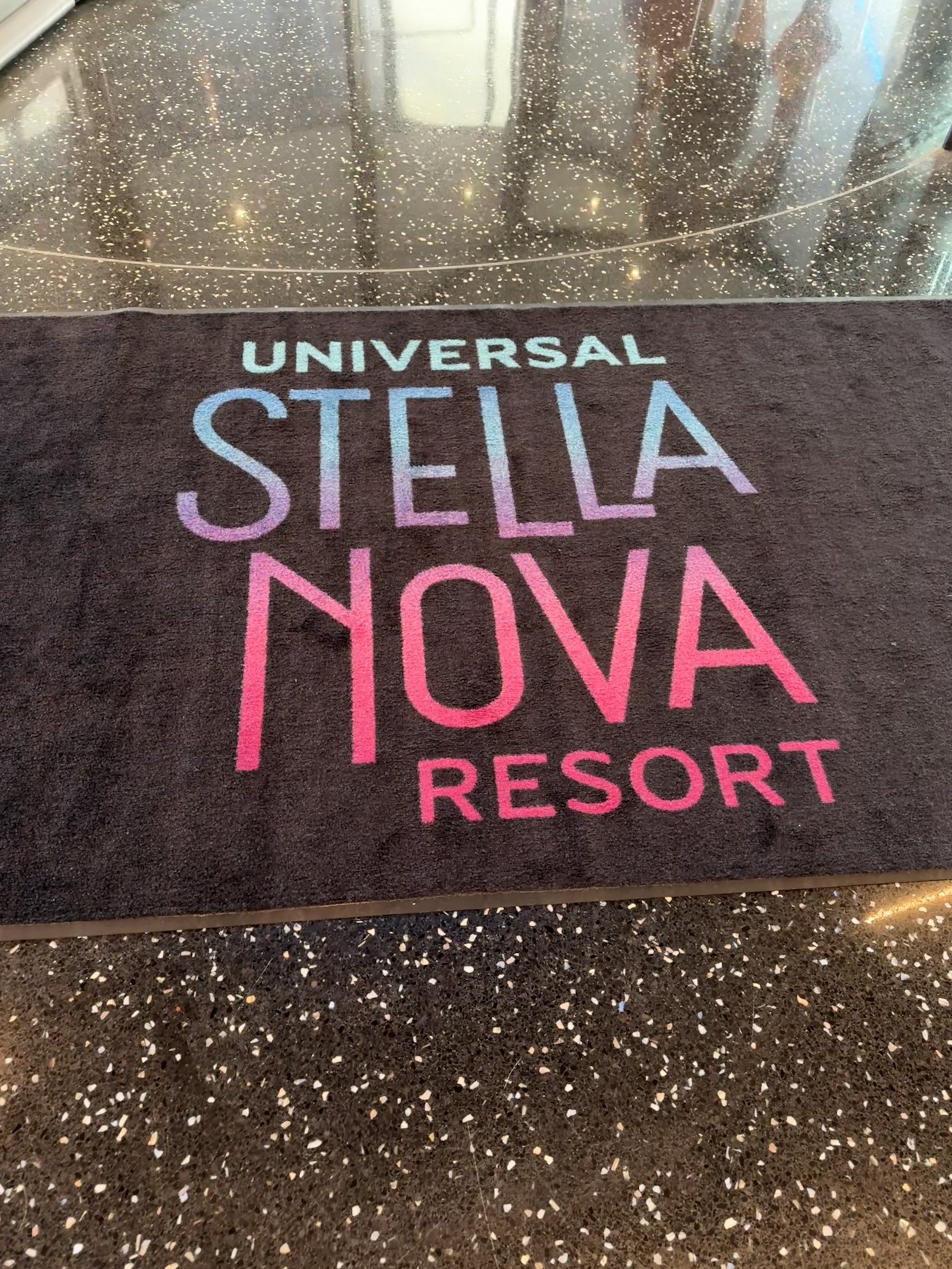 A hotel or resort doormat with the text 'Universal Stella Nova Resort' in colorful gradient letters on a dark background, placed on a speckled floor near glass doors with reflections and a starry pattern.