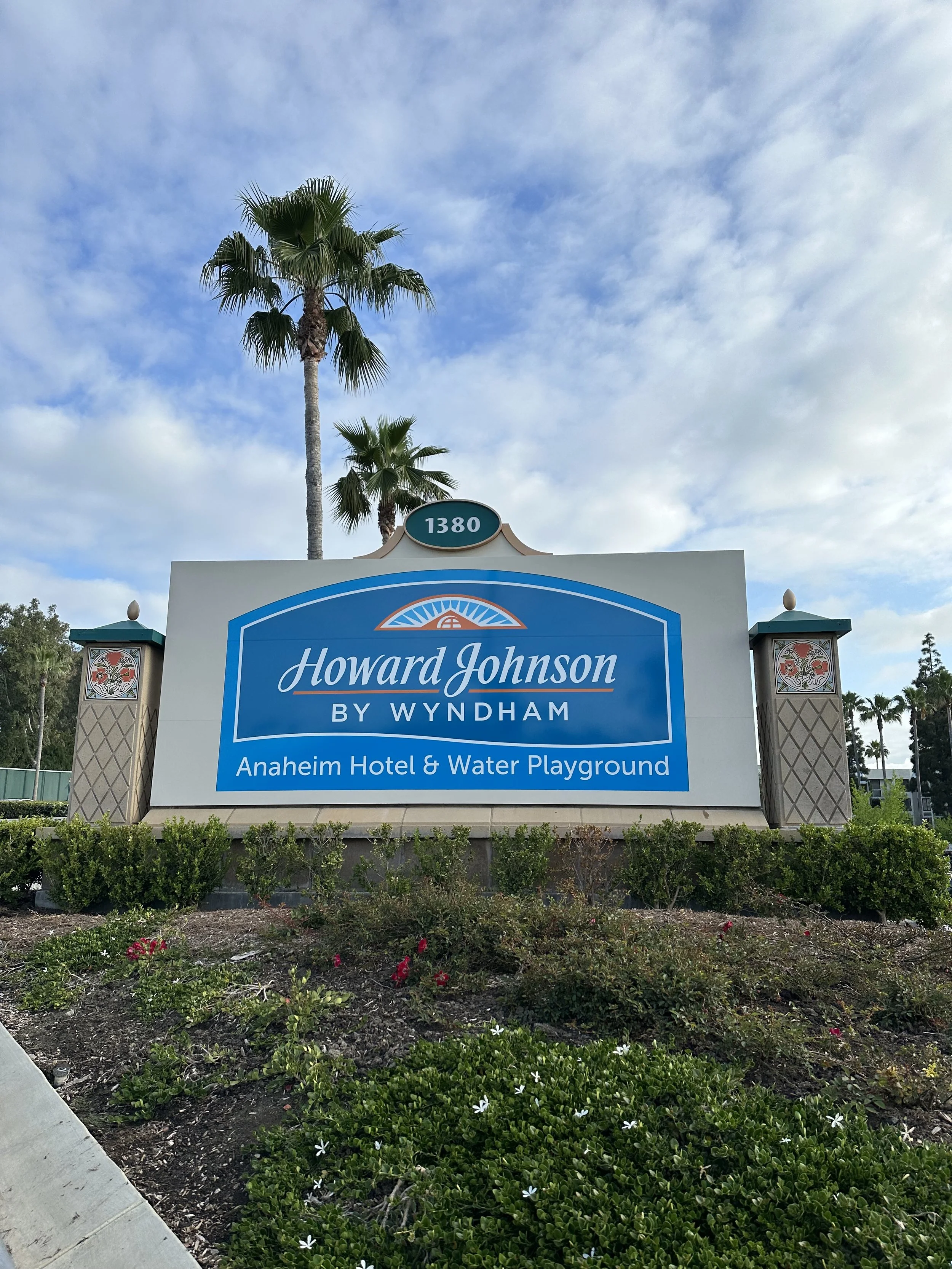 Sign for Howard Johnson by Wyndham Anaheim Hotel & Water Playground, with palm trees and a partly cloudy sky in the background.