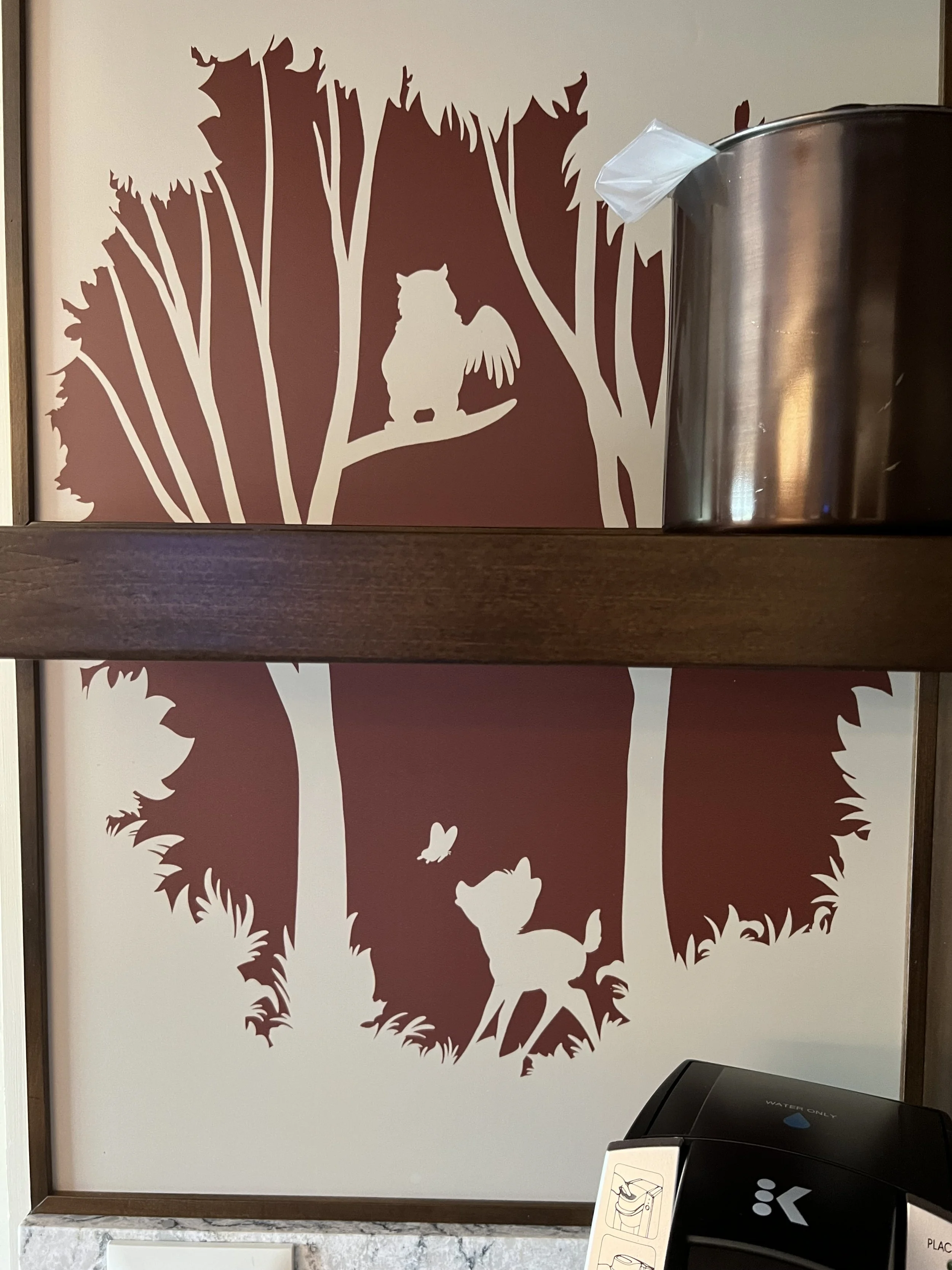 Decorative wall with a forest scene silhouette of a winged owl sitting on a branch and a young deer with butterflies flying around, with a tissue box and water dispenser in front.