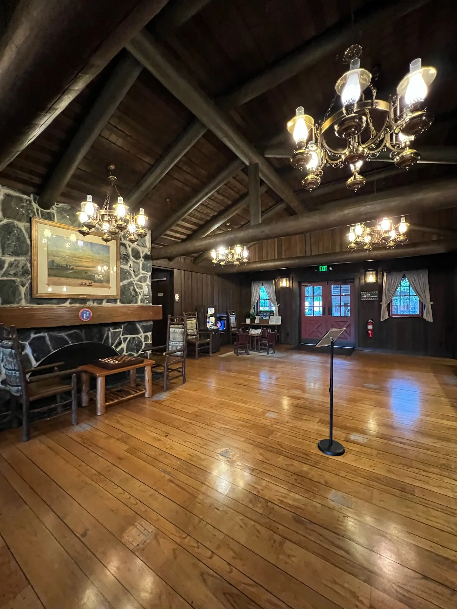Interior of a rustic wooden room with chandeliers, a stone fireplace, chairs, and gaming machines, with polished wooden floors and double doors at the back.